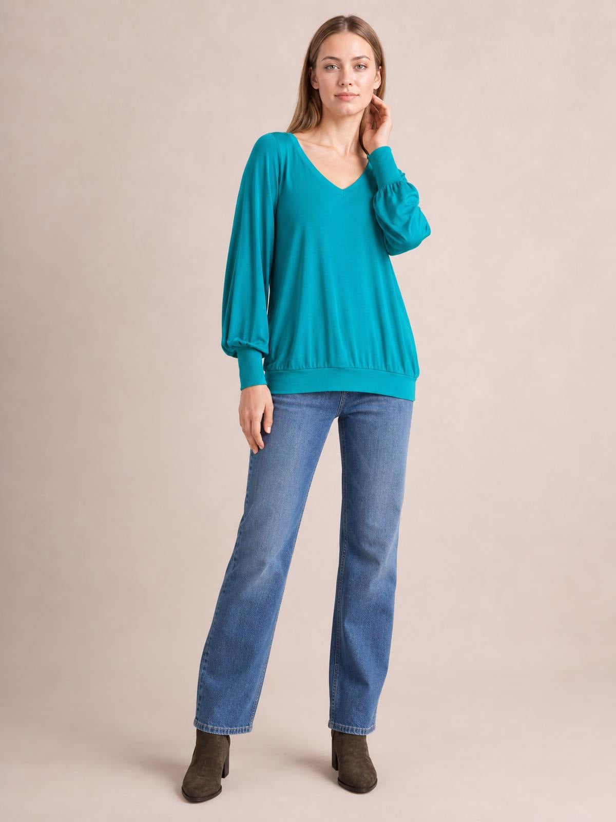 A model poses in a teal V-neck lantern sleeve top with a relaxed silhouette, gathered cuffs, and a wide banded hem.