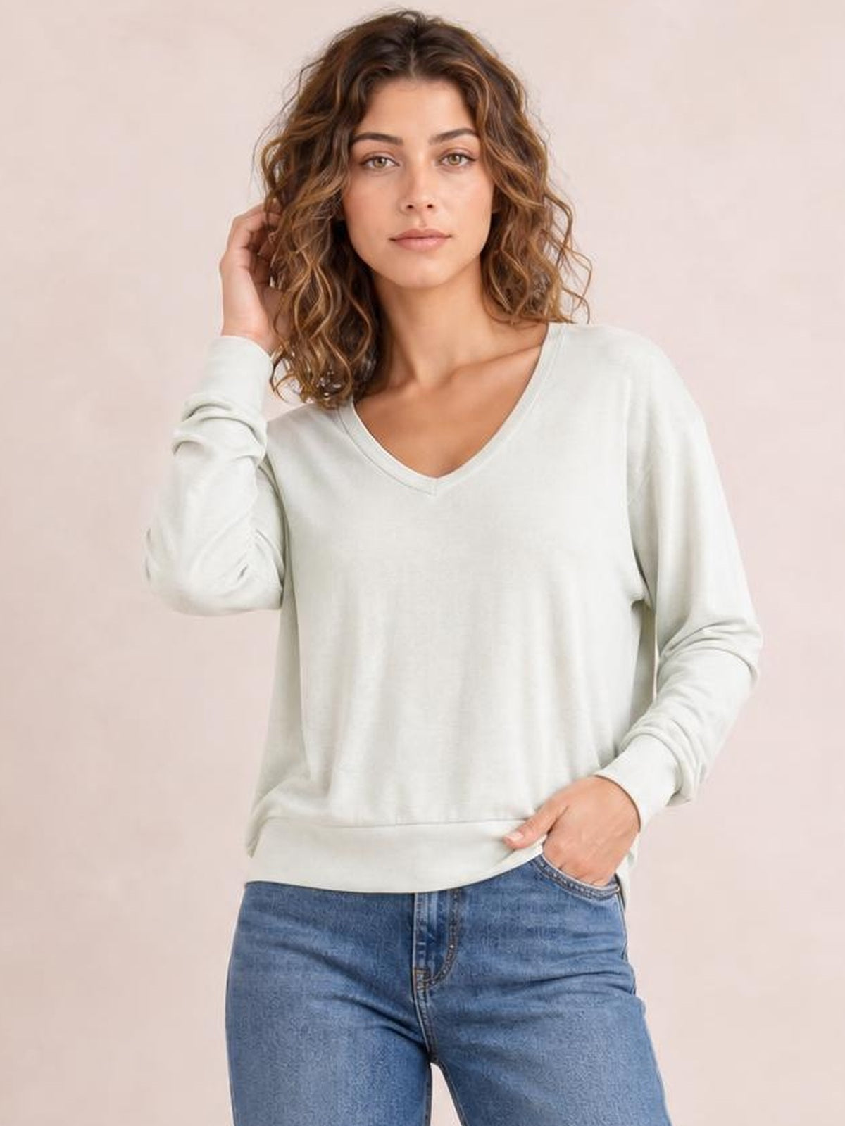 Model wearing an ivory V-neck lantern sleeve top with a relaxed silhouette, long sleeves, ribbed cuffs and hem, posing with one hand in her jeans pocket.