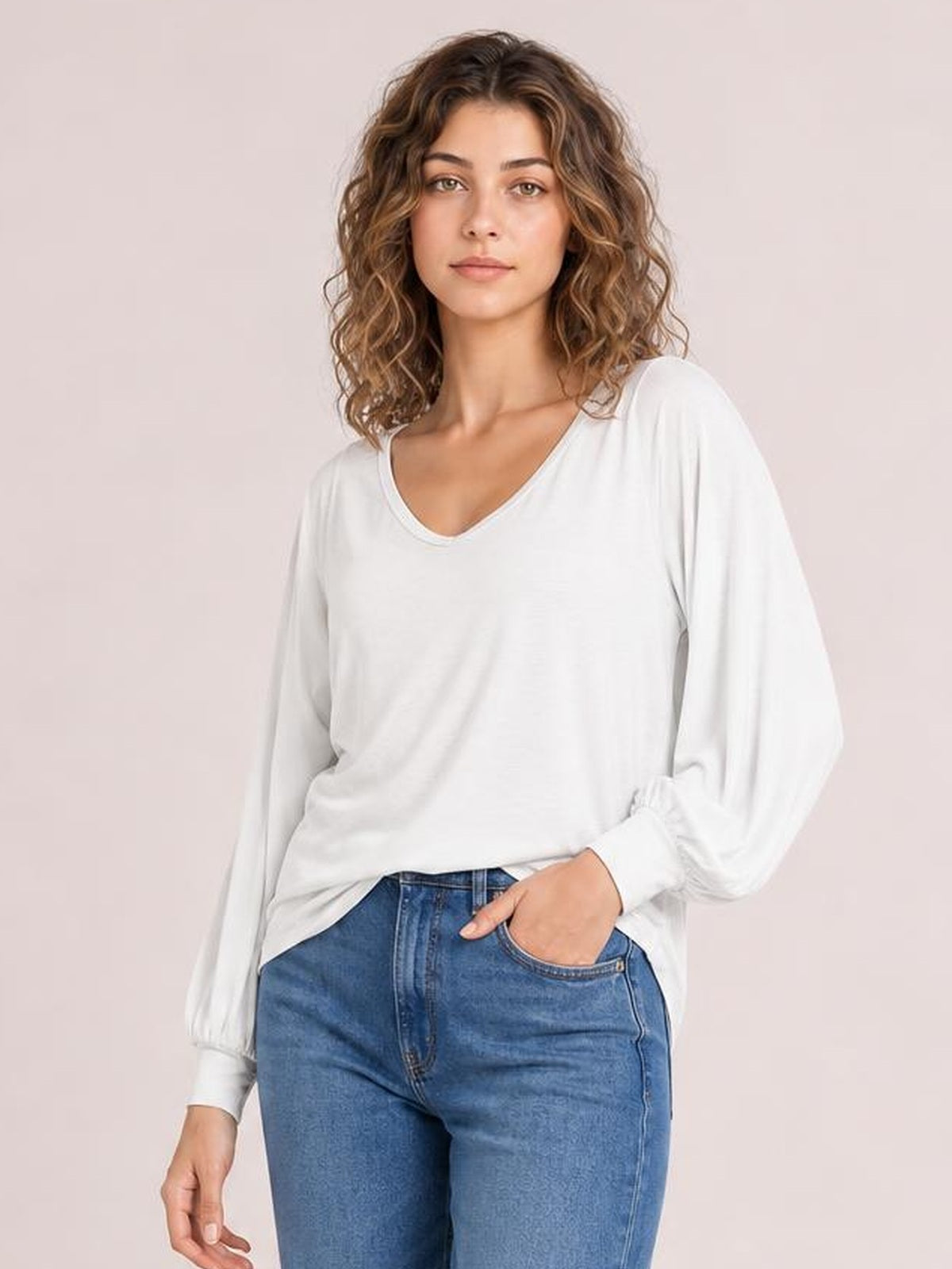 A woman poses in a white V-neck lantern sleeve top with a relaxed silhouette, billowy sleeves, and gathered cuffs, one hand resting in her jeans pocket.