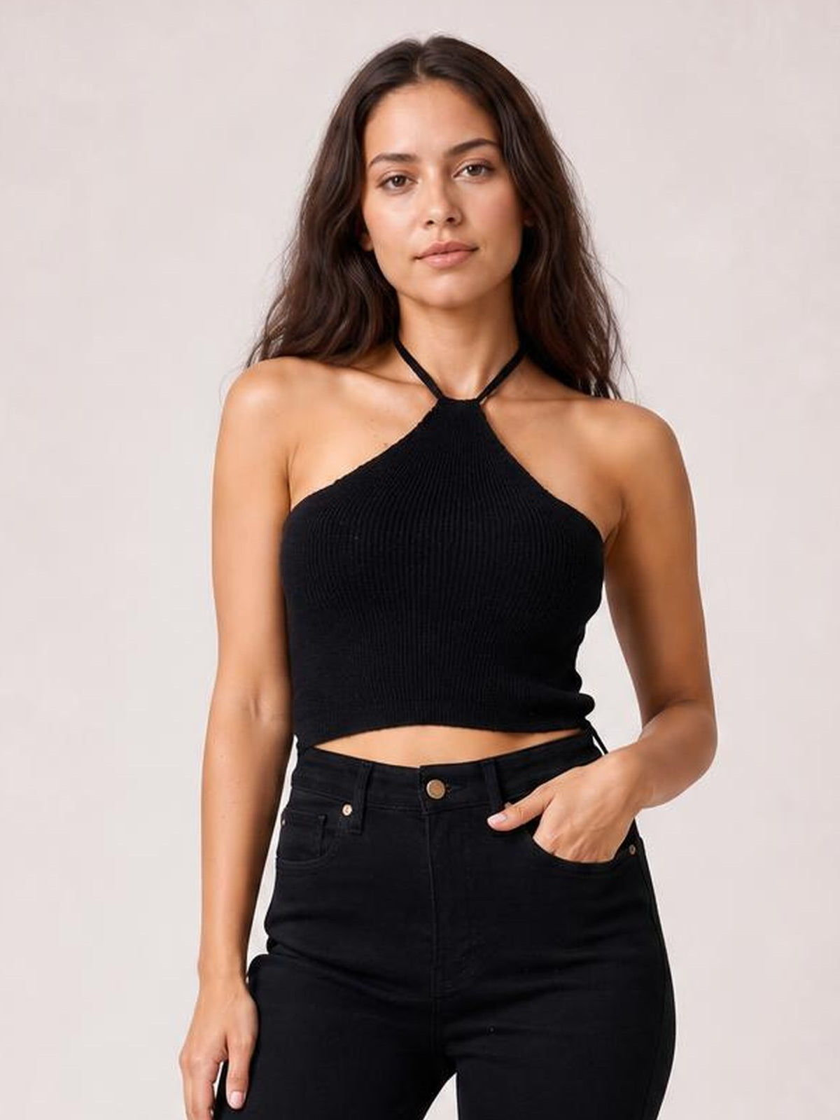 Model wearing a black halter neck ribbed cropped knit top with a fitted silhouette and slim tie straps, paired with high-waisted jeans.