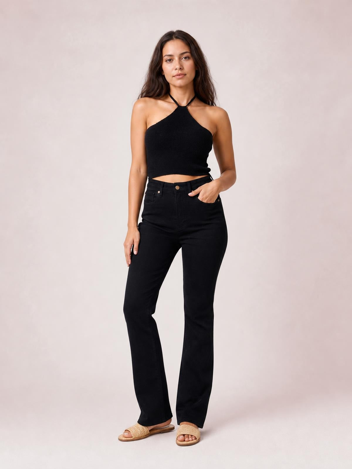 A model poses in a black halter neck ribbed cropped knit top with a tied neckline and fitted silhouette, paired with high-waisted black jeans.