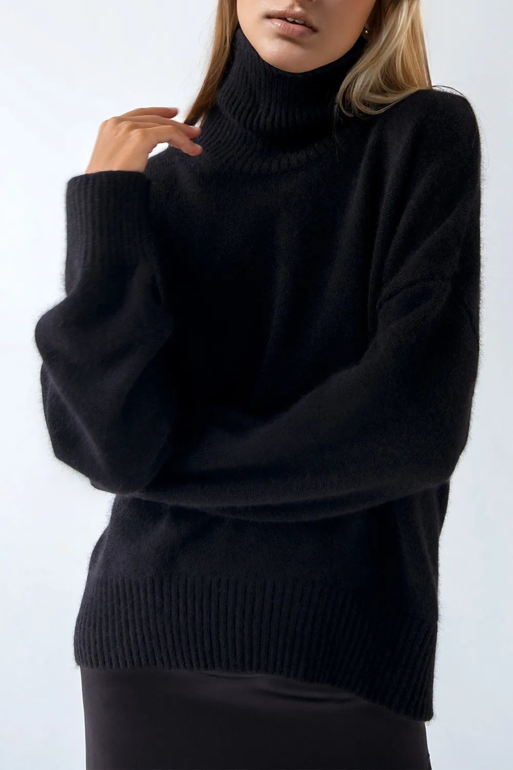 Model wears a black turtleneck dropped-shoulder long sleeve sweater with a relaxed silhouette, ribbed collar, cuffs and hem, touching the neckline.