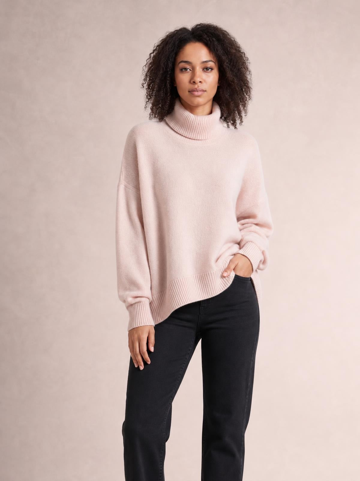 Model wearing a blush pink turtleneck dropped shoulder long sleeve sweater with a relaxed silhouette, ribbed cuffs and hem, and a high-low hemline.