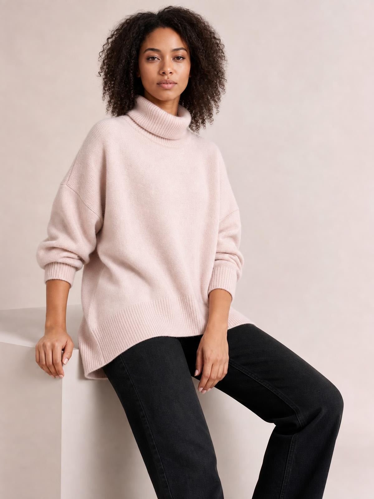 Model seated in a soft blush turtleneck sweater with dropped shoulders, long sleeves, ribbed cuffs and hem, and a relaxed oversized silhouette.