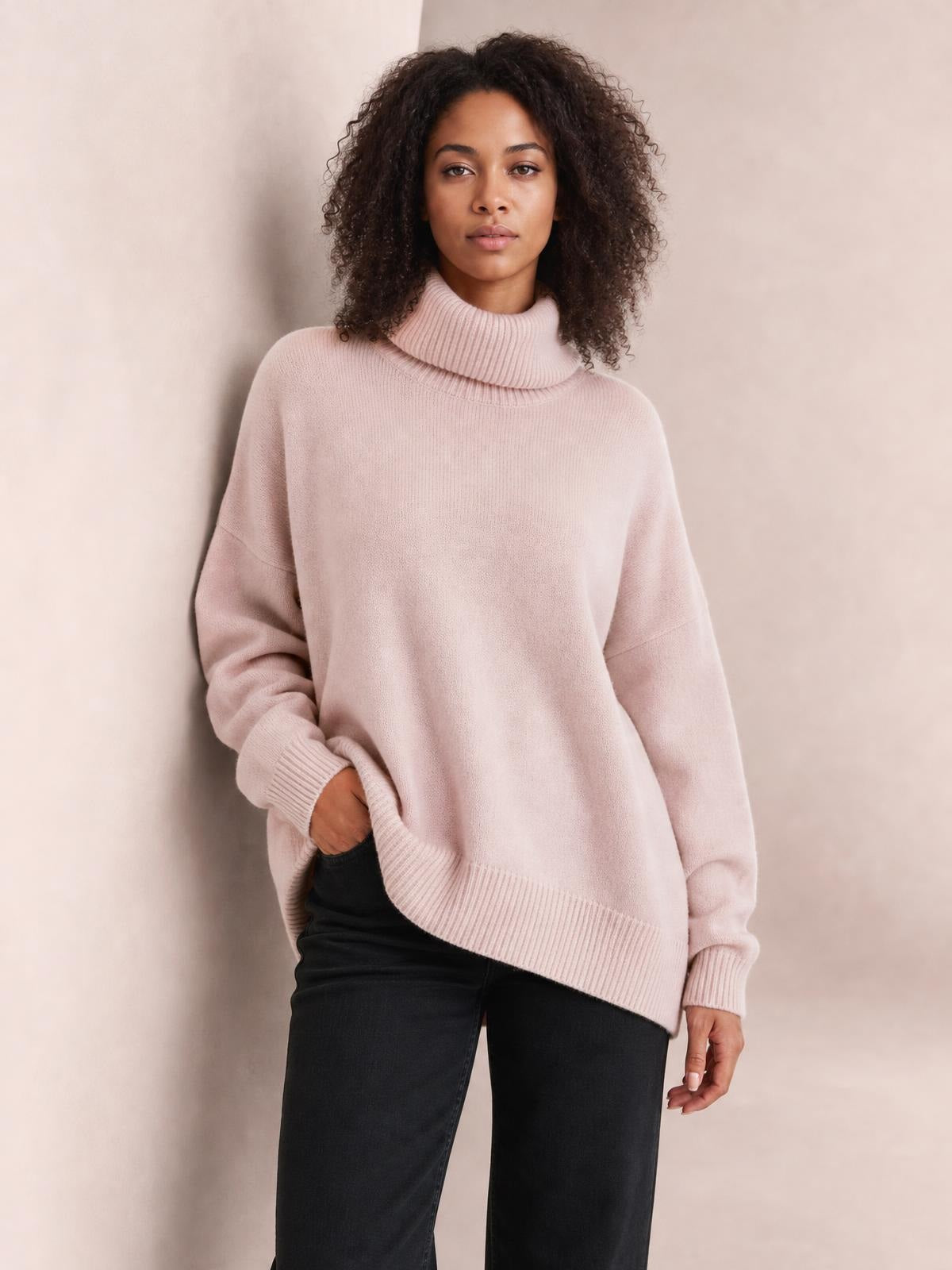 Model wearing a blush pink turtleneck dropped shoulder long sleeve sweater with a relaxed oversized silhouette, ribbed cuffs and hem, standing with one hand in her pocket.