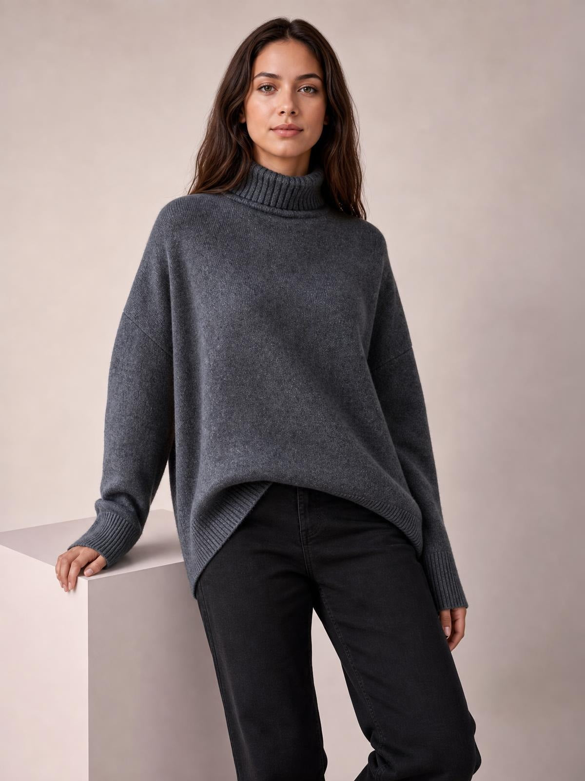 A model leans on a pedestal wearing a charcoal gray turtleneck sweater with dropped shoulders, long sleeves, ribbed cuffs and hem, and a relaxed oversized silhouette.