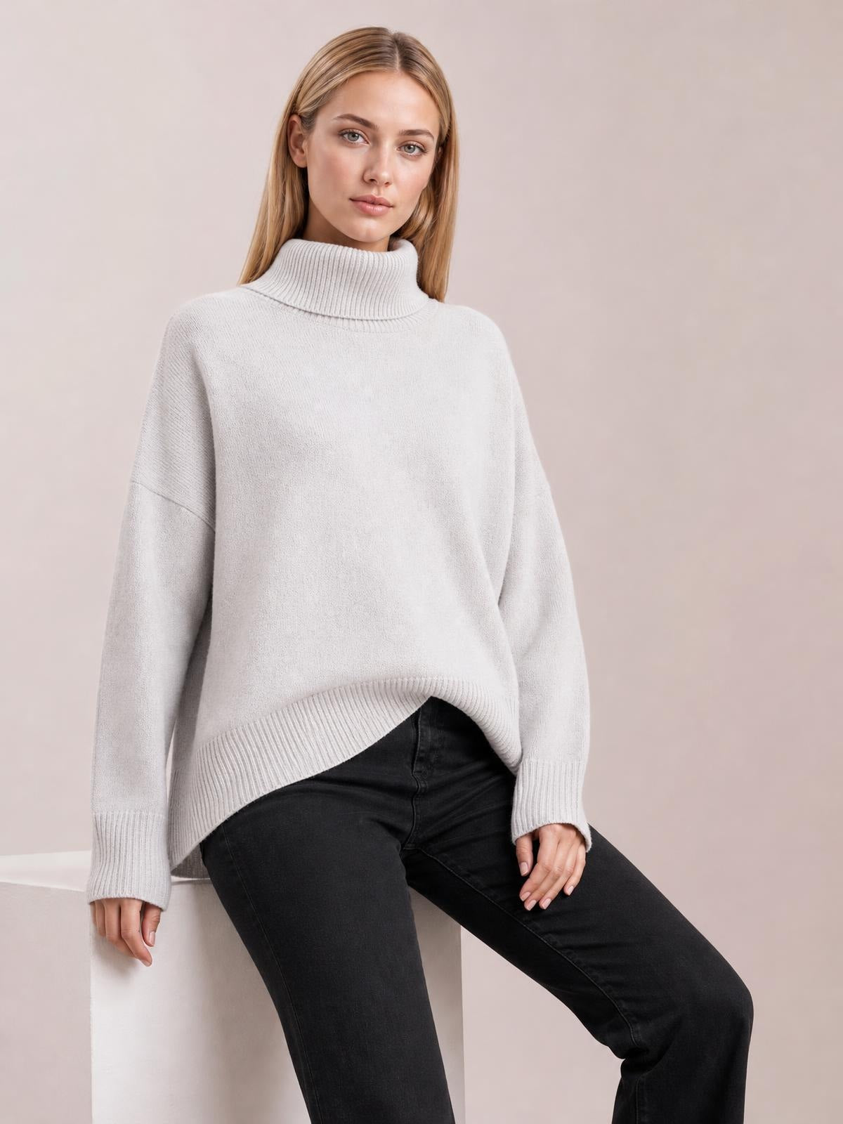 Model seated in a relaxed pose wears a light gray turtleneck dropped-shoulder long-sleeve sweater with a loose silhouette and ribbed cuffs and hem.