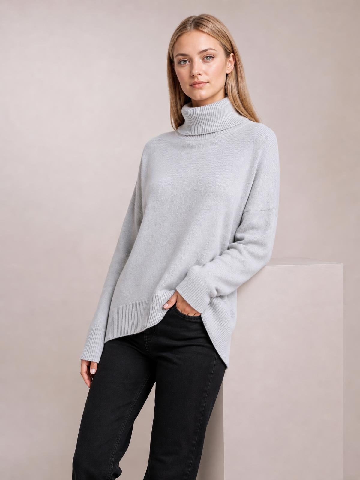 Model wears a light gray turtleneck dropped-shoulder long-sleeve sweater with a relaxed silhouette, ribbed cuffs and hem, posing with one hand in a pocket.