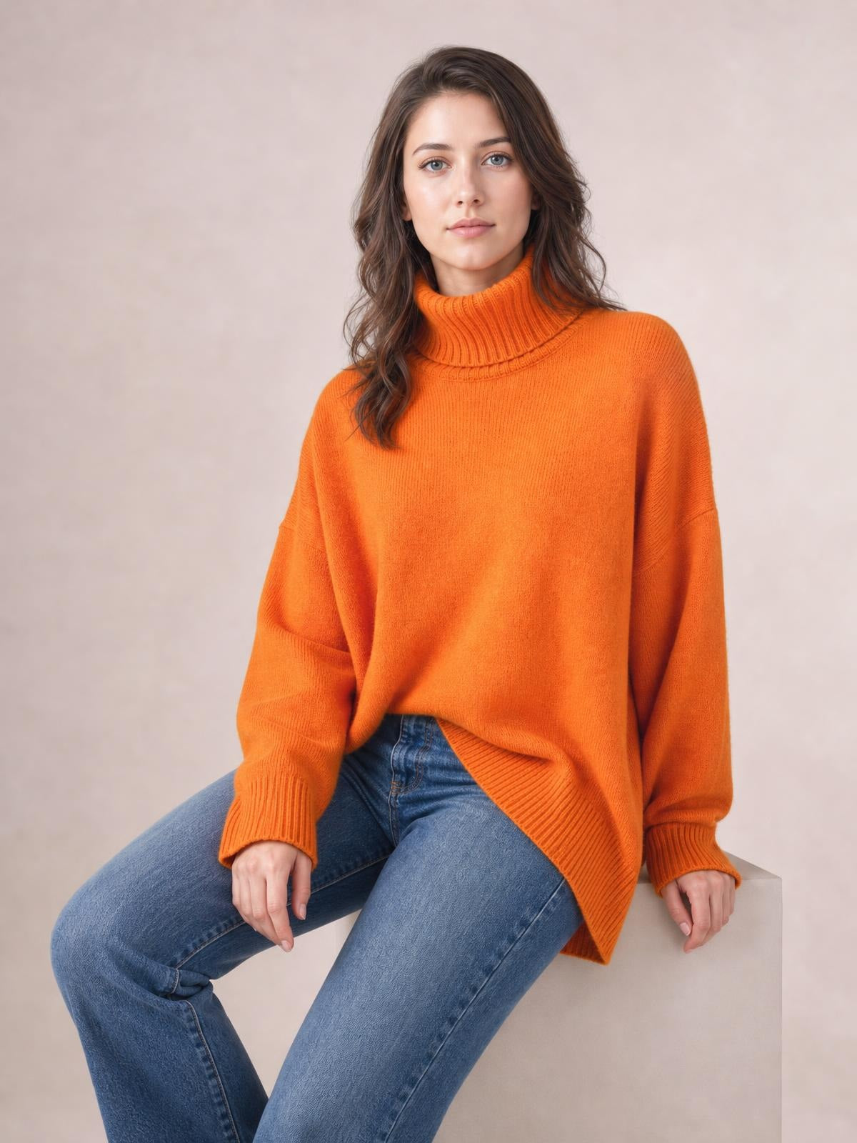 Model seated in an orange turtleneck sweater with dropped shoulders, long sleeves, ribbed cuffs and hem, and a relaxed oversized silhouette.
