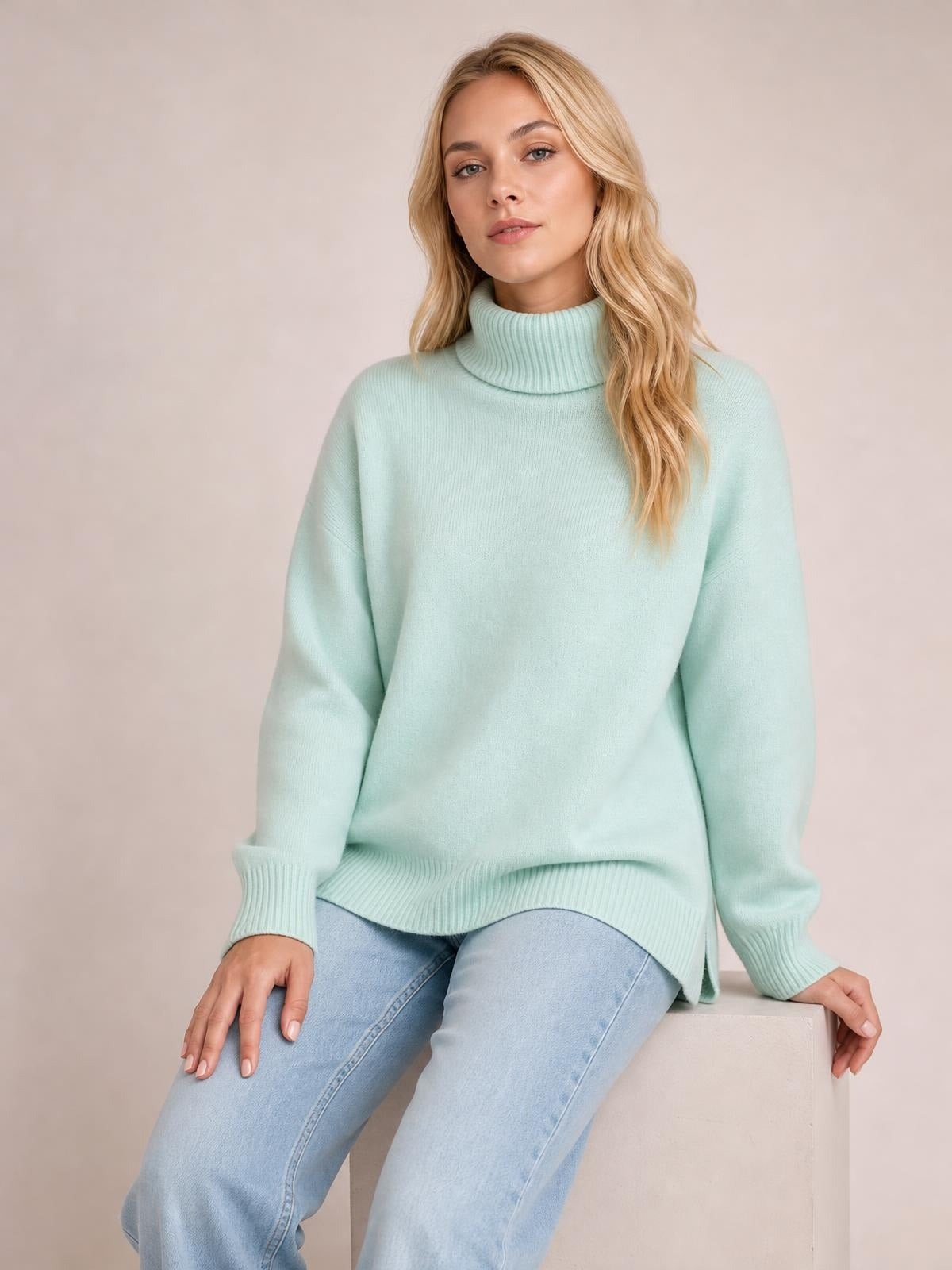 Model seated in a mint green turtleneck dropped-shoulder sweater with long sleeves, ribbed cuffs and hem, and a relaxed oversized silhouette.