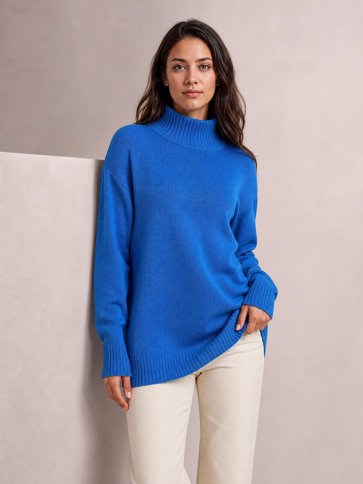 A model wears a bright blue turtleneck sweater with dropped shoulders, long sleeves, ribbed cuffs and hem, and a relaxed silhouette while lifting the hem.
