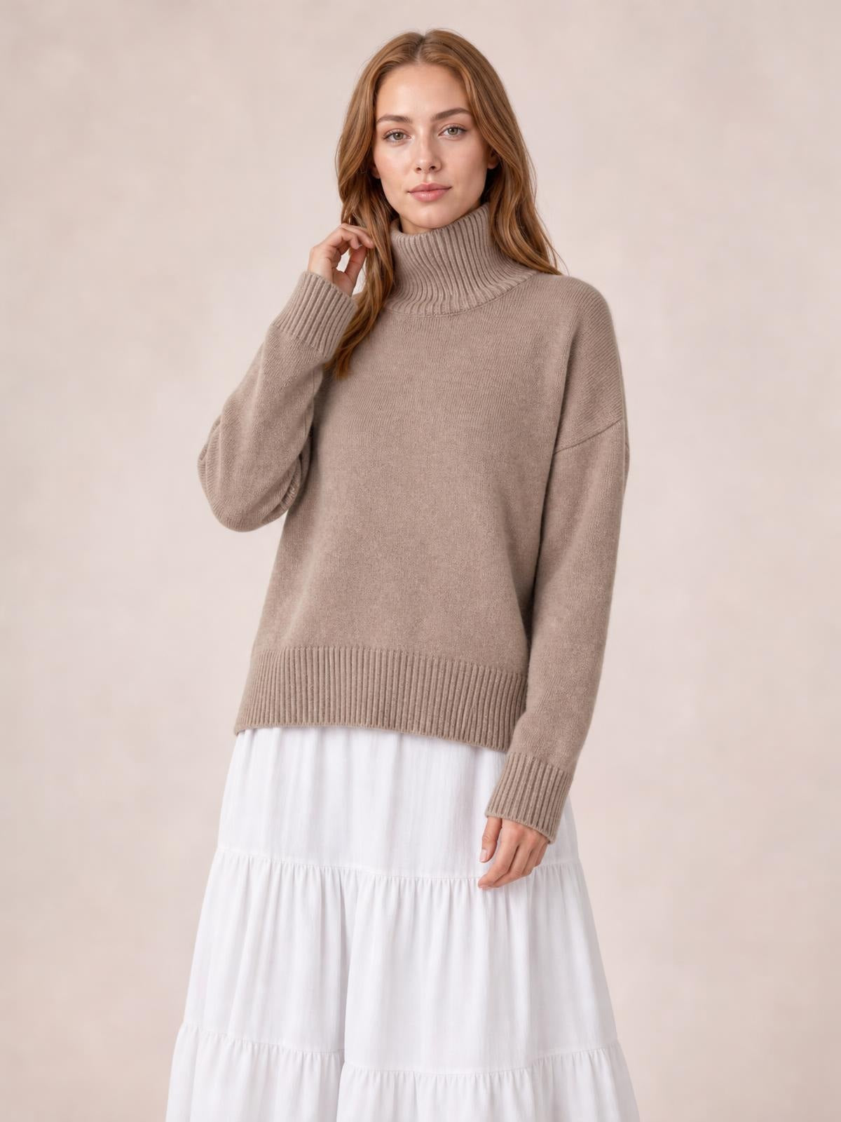 Model wears a taupe turtleneck dropped-shoulder long-sleeve sweater with a relaxed silhouette, ribbed collar, cuffs, and hem, posing with one hand near her hair.