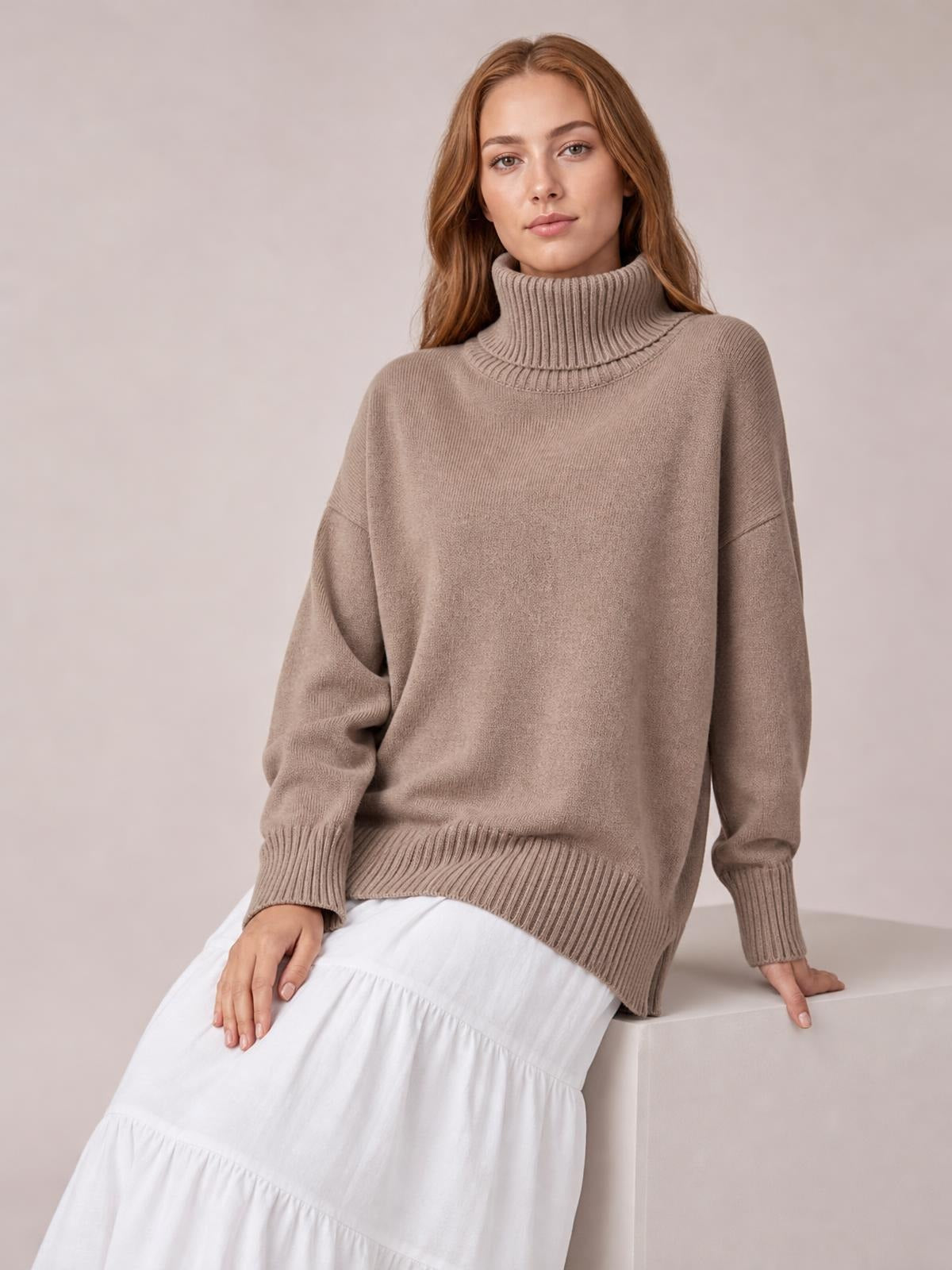 A model sits wearing a taupe turtleneck sweater with dropped shoulders, long sleeves, ribbed cuffs and hem, and a relaxed silhouette.