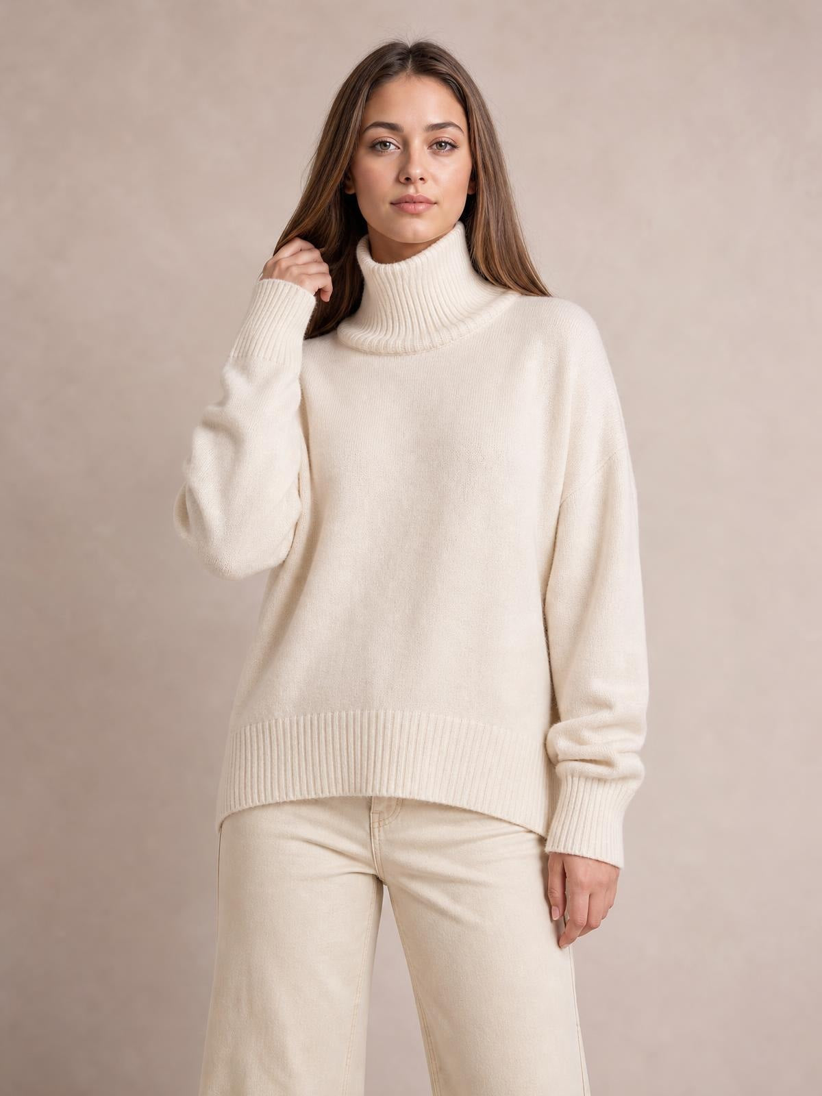 Model wearing a cream turtleneck dropped-shoulder long-sleeve sweater with a relaxed silhouette, ribbed collar, cuffs, and hem, posing with one hand in her hair.