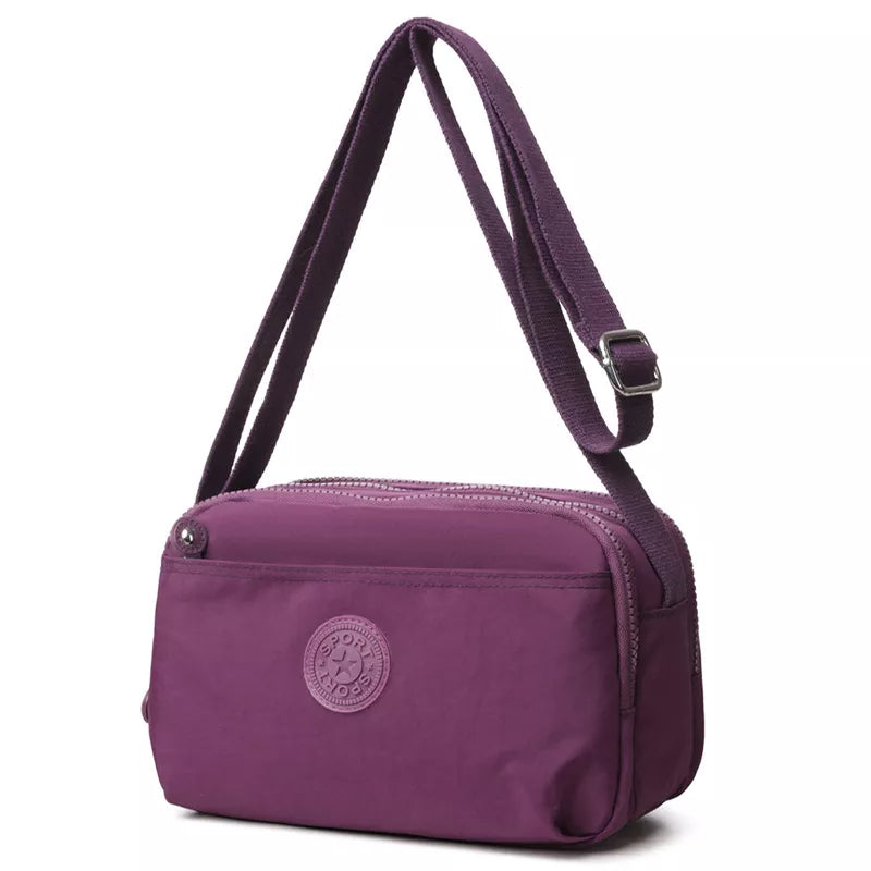 Small purple nylon crossbody bag with an adjustable strap, silver buckle, zip-top closure, rounded silhouette, and front zip pocket with logo patch.