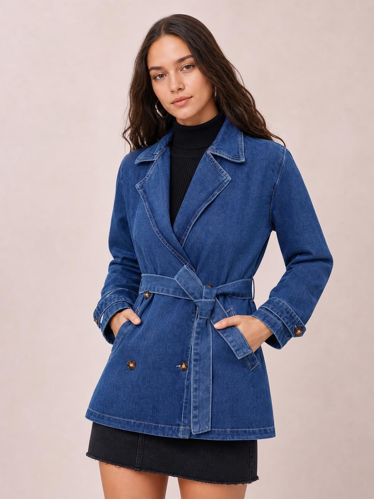 A model stands with hands in the pockets of a blue belted double-breasted denim jacket featuring notched lapels, long sleeves, button details, and a relaxed silhouette.