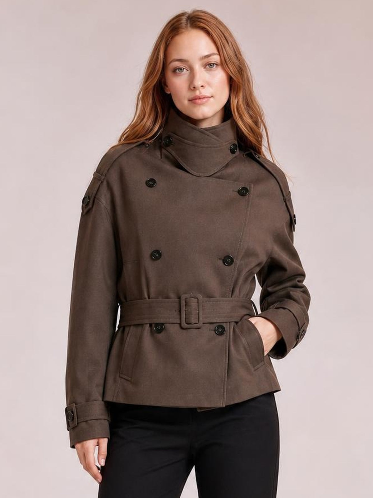 Model poses with one hand in a pocket wearing a relaxed brown double-breasted trench jacket with button details, wide belt, and cuff straps.