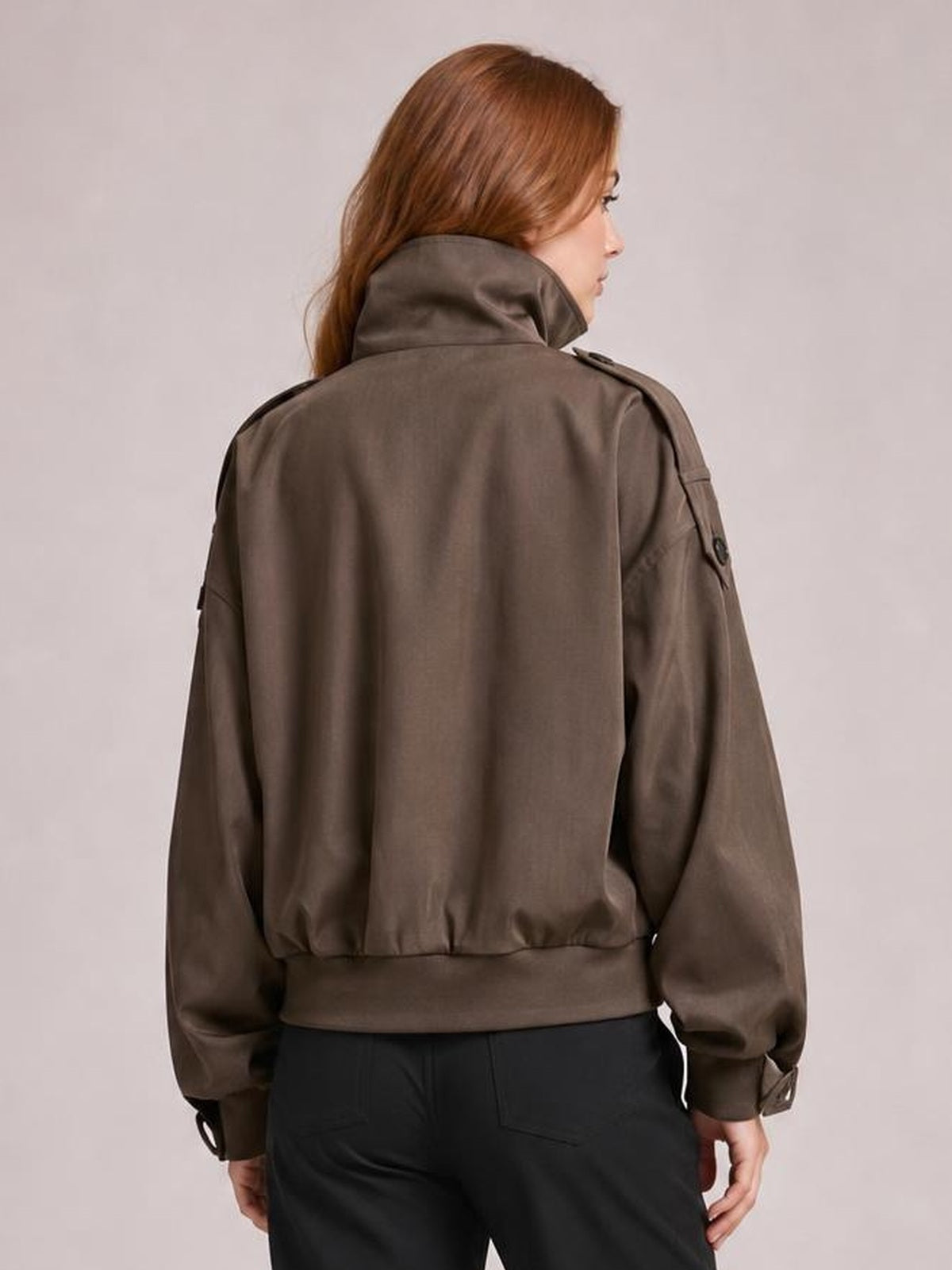 A model shown from behind wears a relaxed taupe-brown trench jacket with a high collar, long sleeves, shoulder tabs, cuff straps, and a gathered hem.
