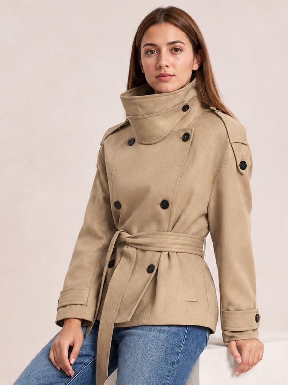 A seated model wears a relaxed-fit beige belted waist button-up trench jacket with a high collar, double-breasted buttons, shoulder tabs, and cuff straps.