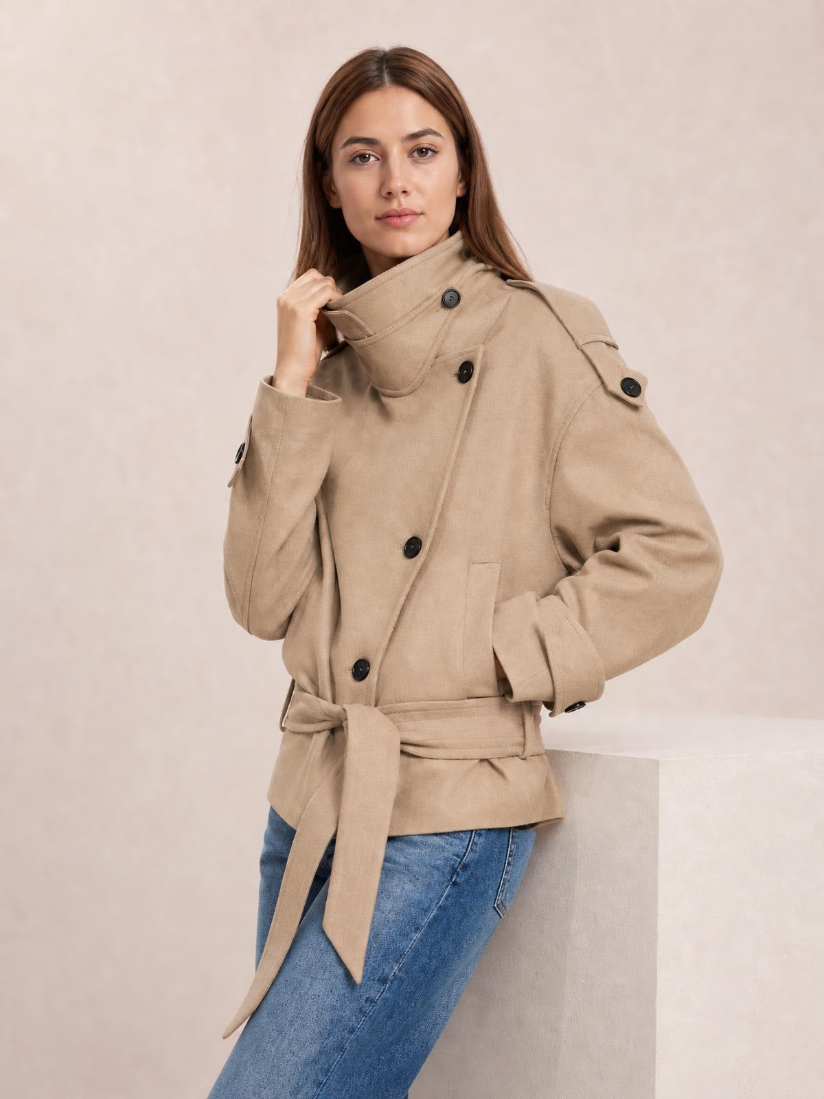Model adjusts the collar of a relaxed-fit beige button-up trench jacket with double-breasted buttons, shoulder tabs, cuff straps, pockets, and a tied waist belt.