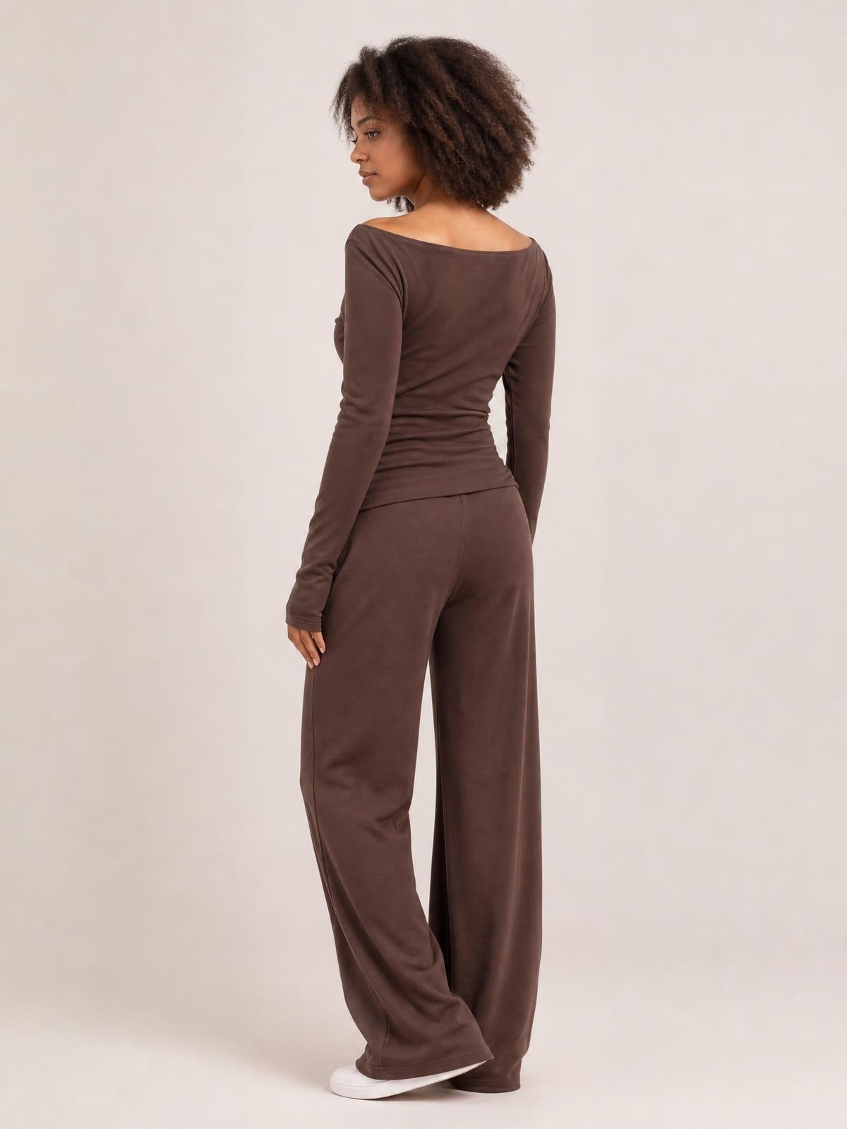 A model poses turned away in a brown boat-neck long-sleeve top and coordinating wide-leg pants, showing the fitted silhouette and relaxed drape.