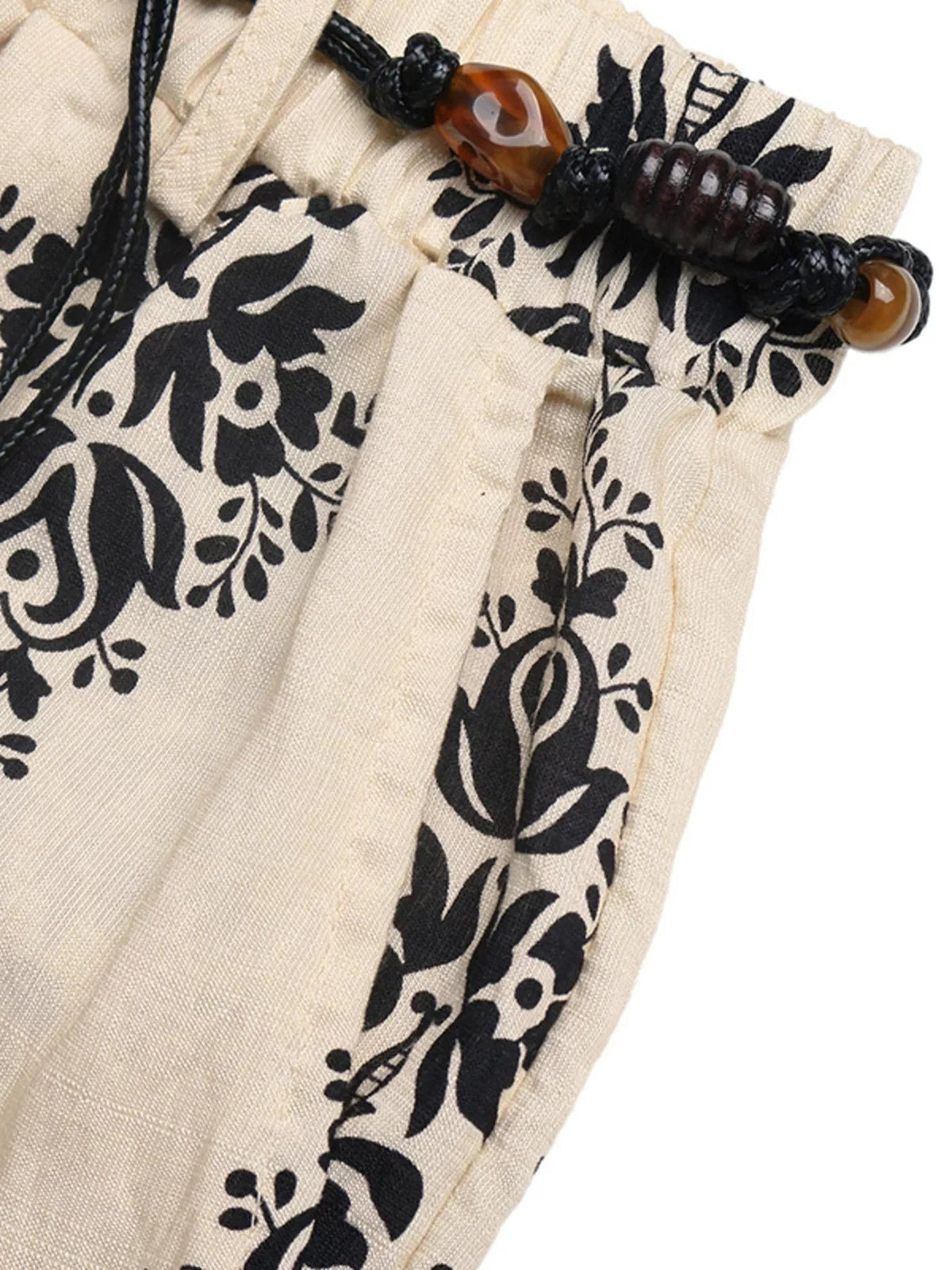 Close-up of cream boho print wide leg pants with black botanical motifs, a braided drawstring waistband, and wood-tone bead accents.