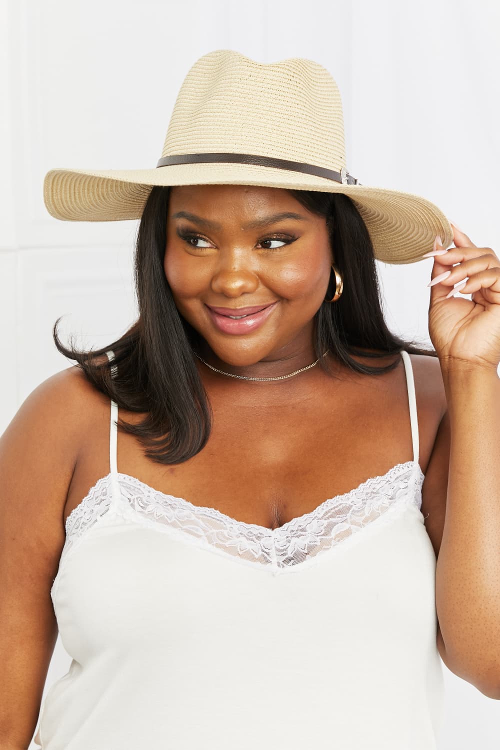Model smiles while holding an ivory boho straw fedora with a softly structured crown, wide brim, woven texture, and contrasting dark buckle band.