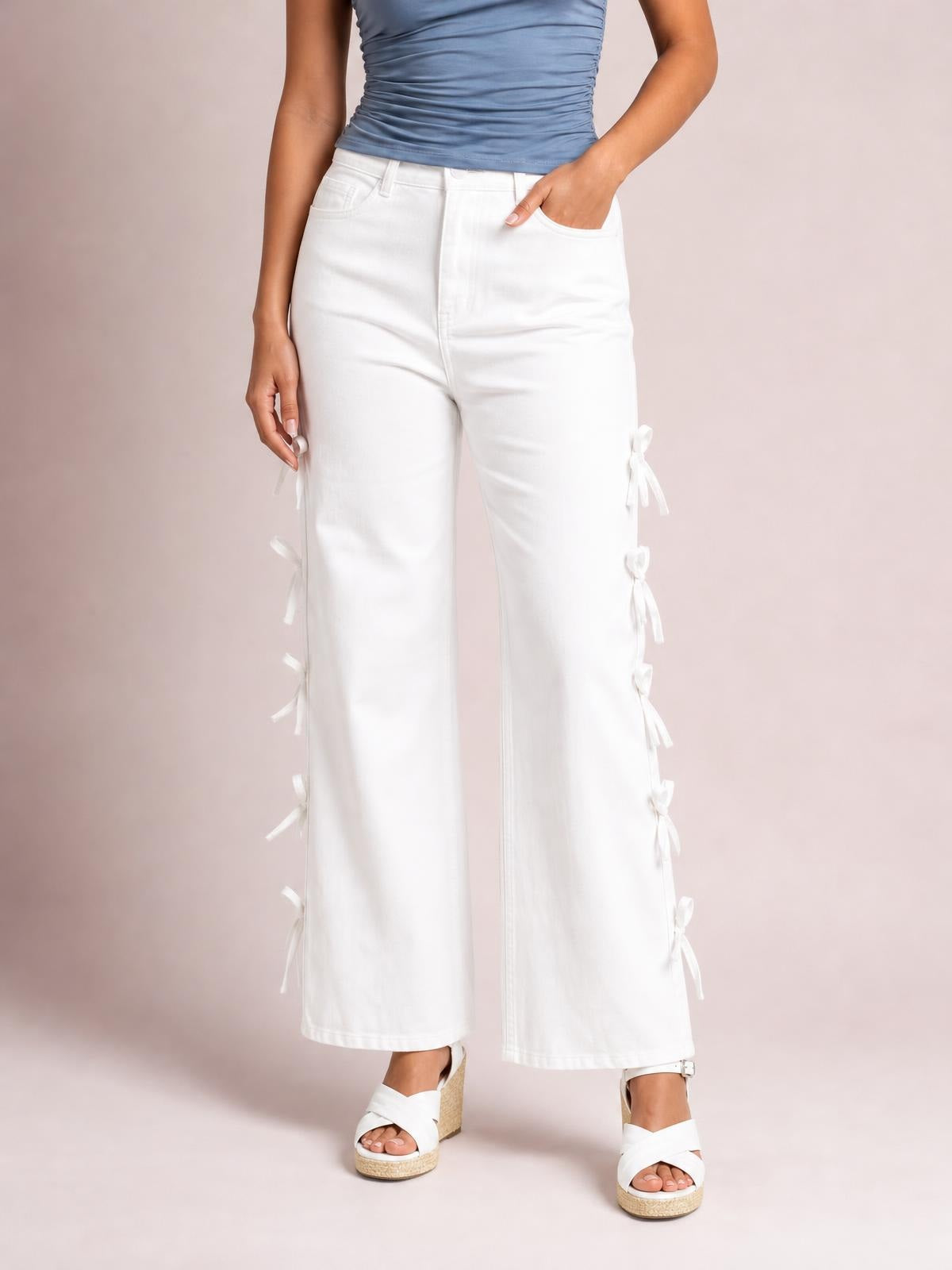 Model wears white wide-leg pants with a structured waistband, front pockets, and multiple bow accents tied along the outer sides.