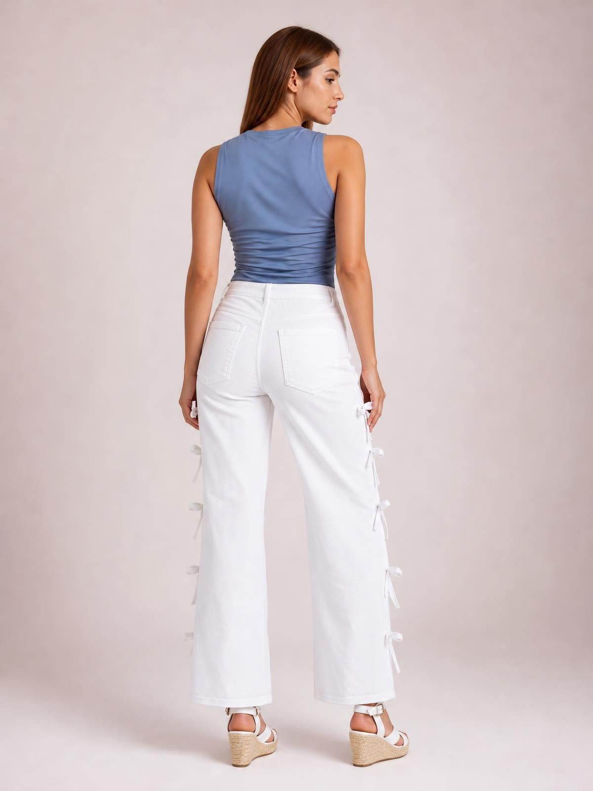 A model poses from the back in white bow-accented wide-leg pants with a high waist, belt loops, back pockets, cropped hems, and side-tie details.