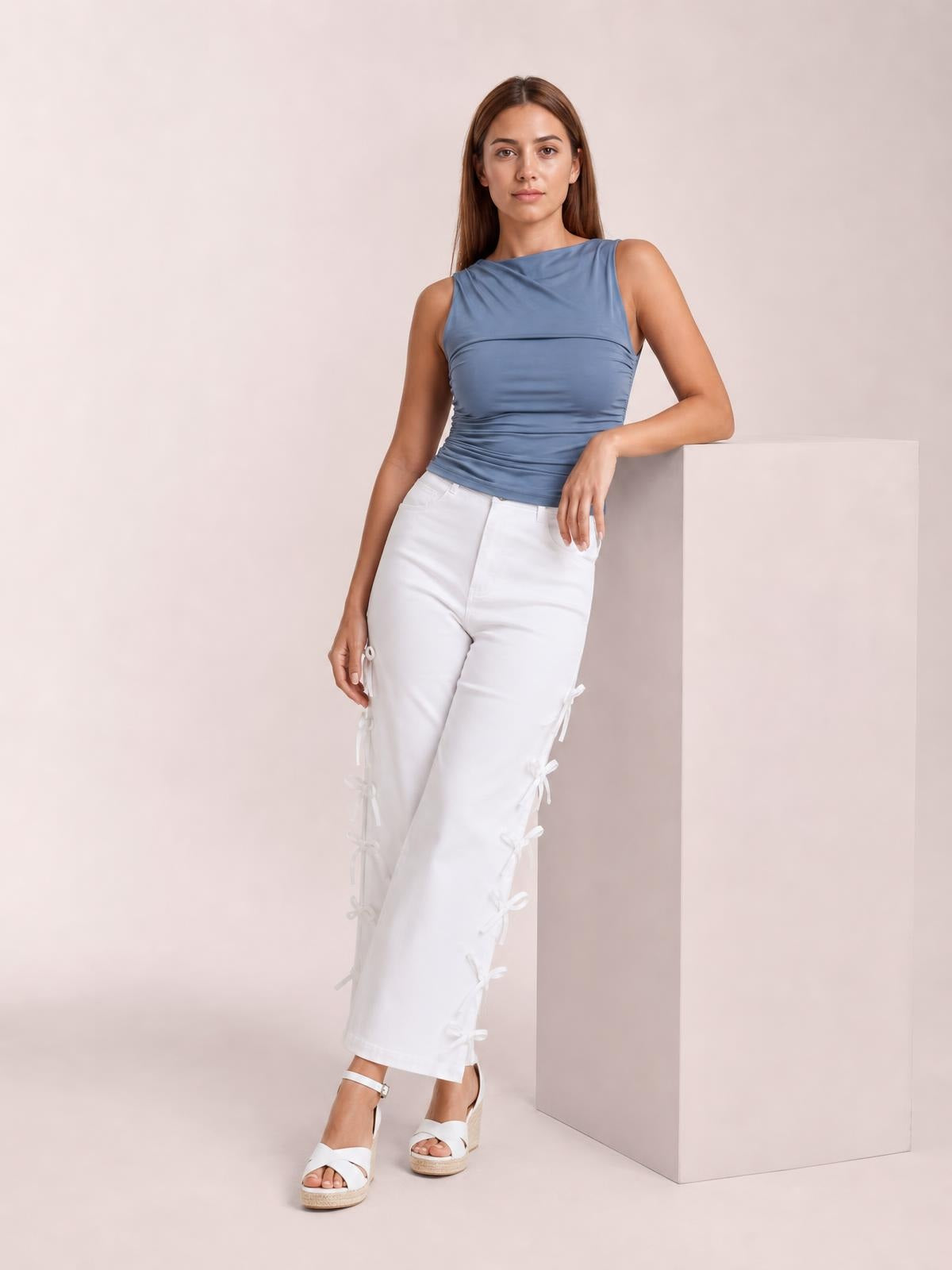 A model leans against a pedestal wearing white bow-accented wide-leg pants with a structured waistband, cropped hems, and side-tie detailing.