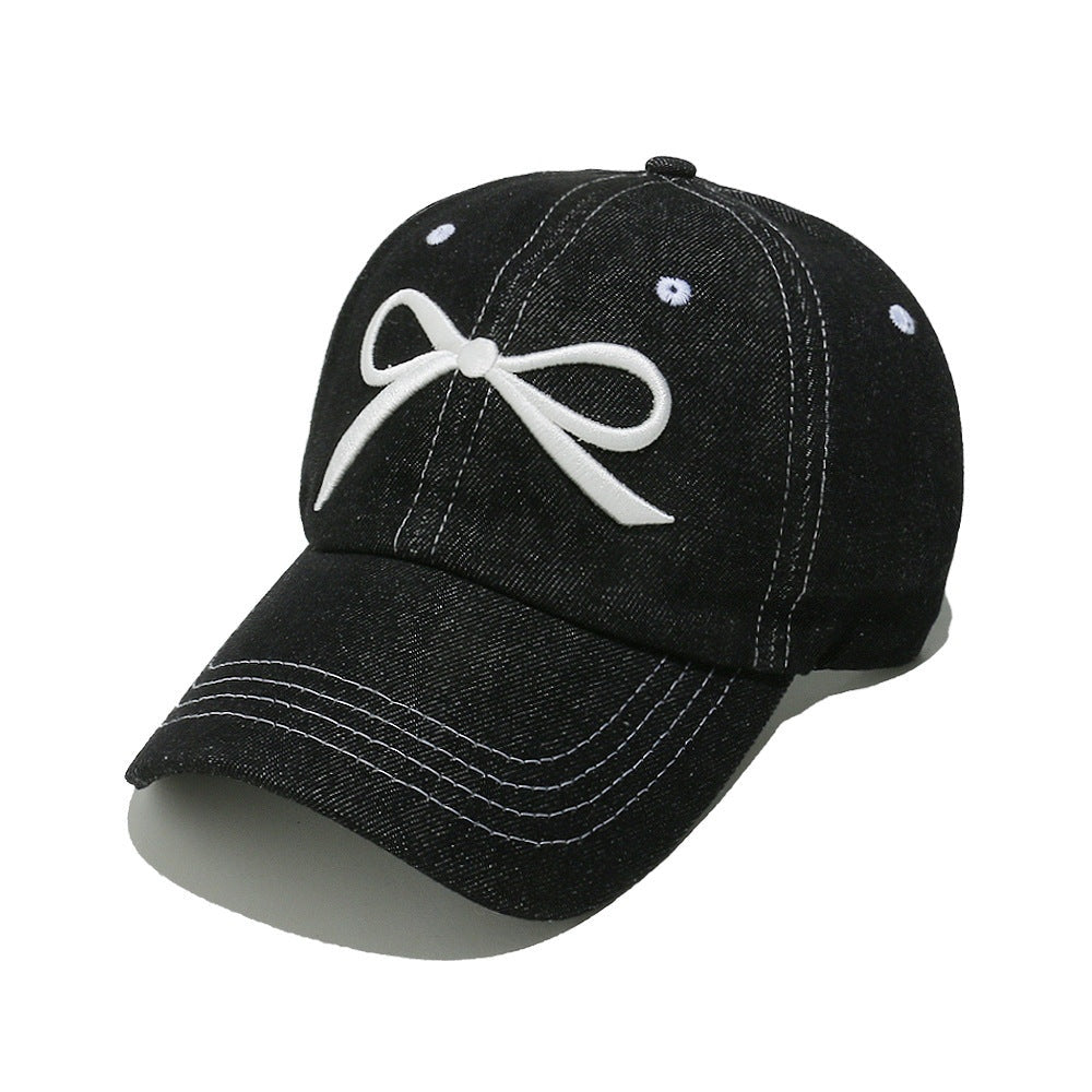 Black denim cotton baseball cap with a white embroidered bow, six-panel crown, curved brim, contrast topstitching, ventilation eyelets, and crown button.