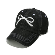 Black denim cotton baseball cap with a white embroidered bow, six-panel crown, curved brim, contrast topstitching, ventilation eyelets, and crown button.