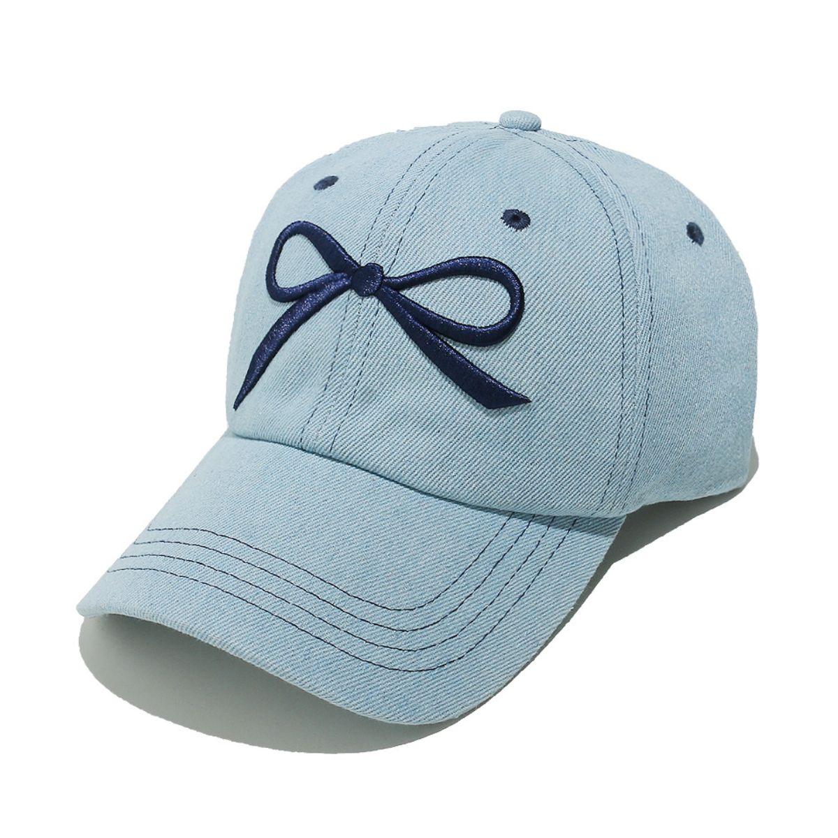 Light blue denim cotton baseball cap with a six-panel silhouette, curved brim, dark embroidered bow, visible topstitching, ventilation eyelets, and crown button.