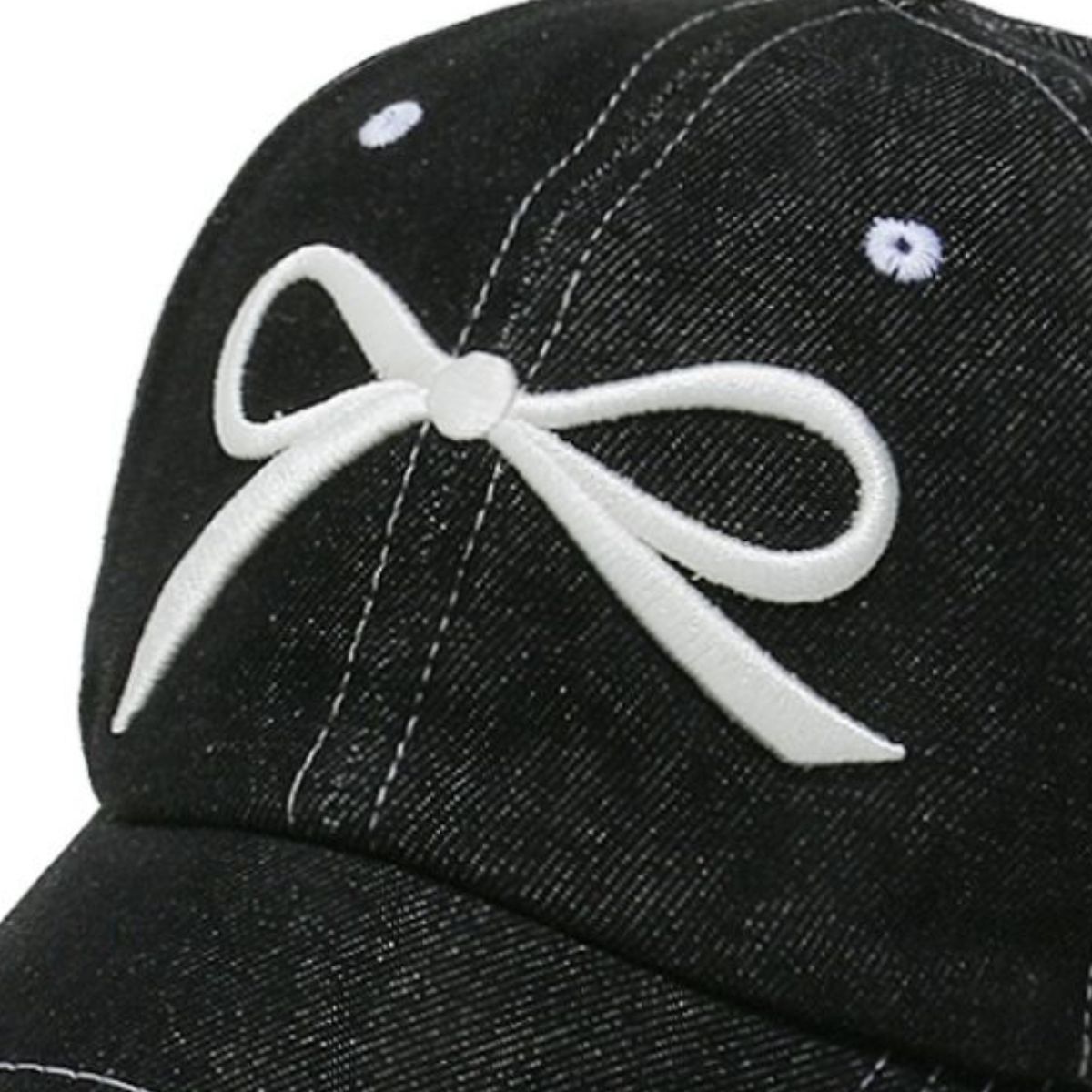 Close-up of a black denim cotton baseball cap featuring a white embroidered bow, six-panel construction, contrast topstitching, ventilation eyelets, and a curved brim.