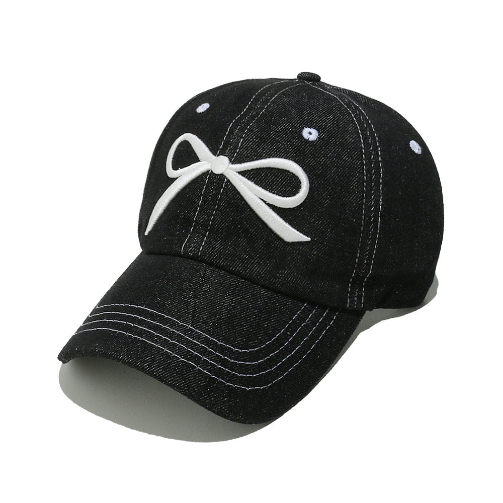 Black denim cotton baseball cap with a white embroidered bow, six-panel crown, curved brim, contrast topstitching, ventilation eyelets, and crown button.