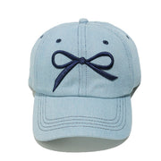Light blue denim cotton baseball cap with a dark embroidered bow, curved brim, six-panel construction, contrast topstitching, ventilation eyelets, and crown button.