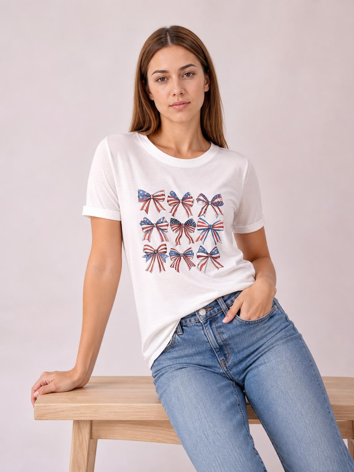 Model seated in a relaxed pose wears a white round-neck short-sleeve T-shirt with a colorful bow graphic print and straight, slightly loose silhouette.