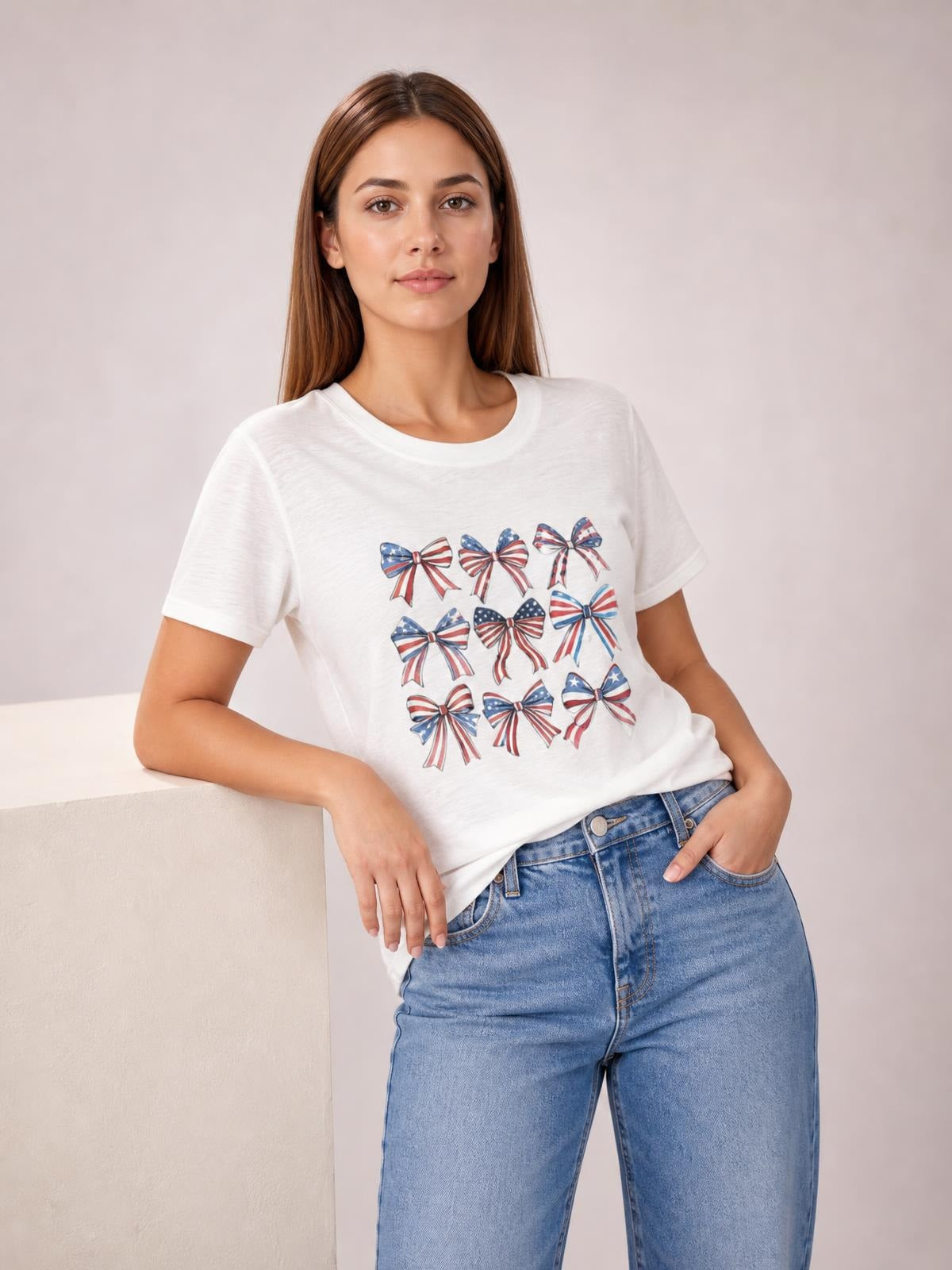 Model leans against a ledge wearing an opaque white round-neck short-sleeve T-shirt with a colorful bow graphic and a relaxed silhouette.