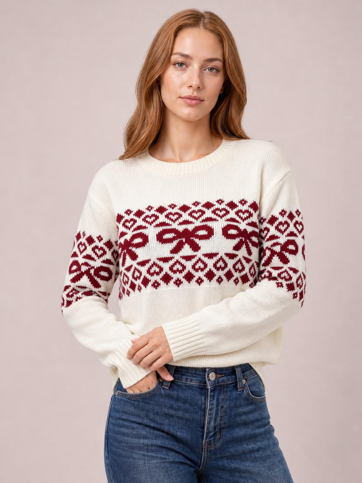 Woman wearing an ivory long-sleeve Christmas sweater with red bow and geometric knit patterns, ribbed crew neck and cuffs, relaxed silhouette, hands near pockets.