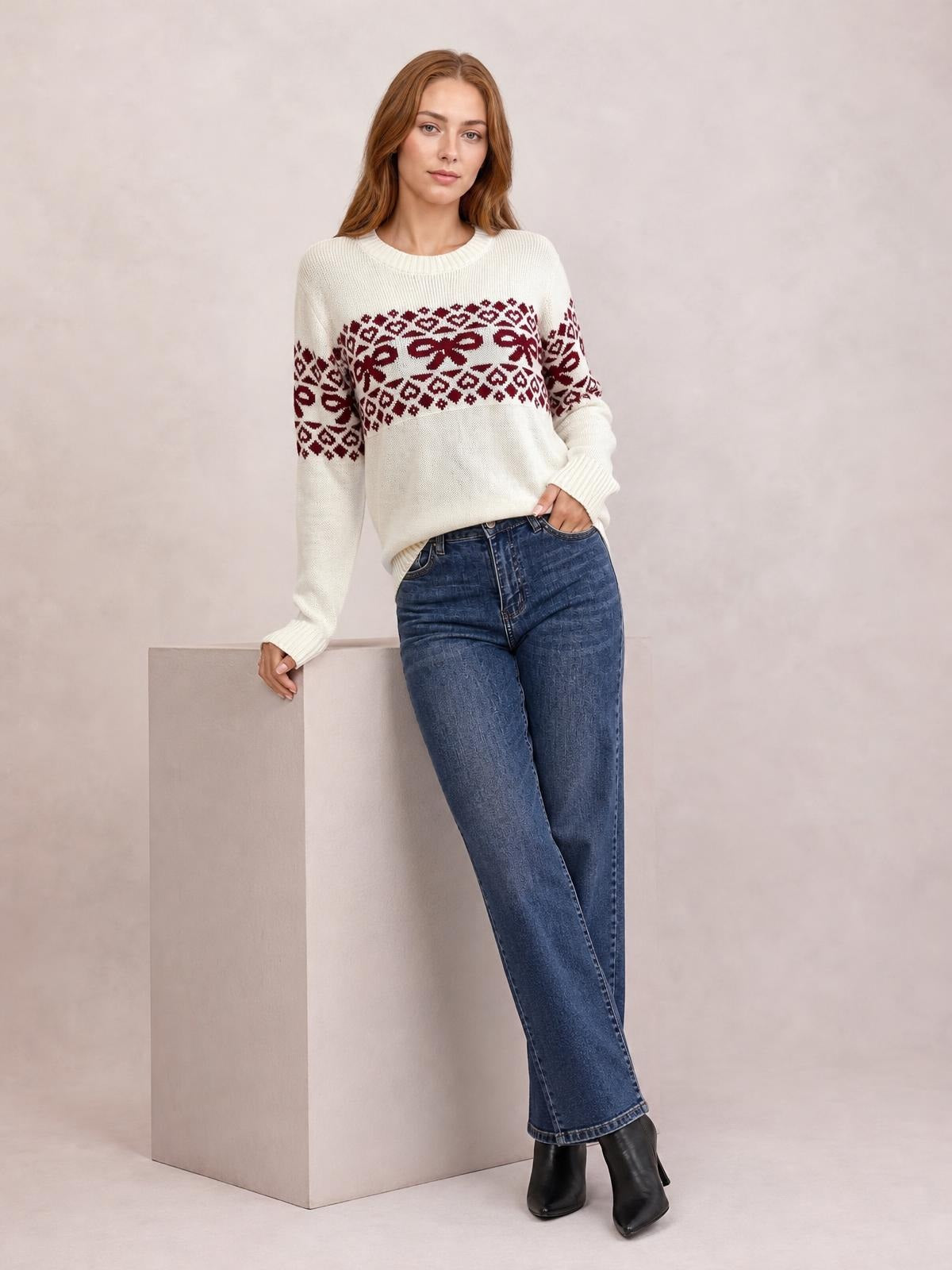 Model wears a cream long-sleeve Christmas sweater with burgundy bow and geometric patterns, ribbed crew neck and cuffs, paired with jeans.
