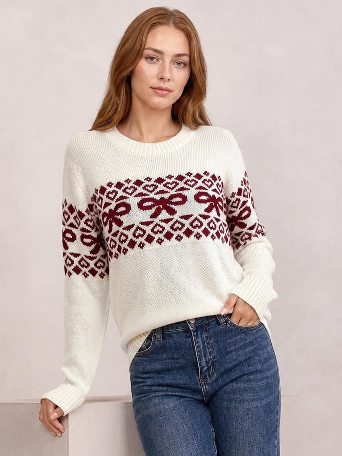 Model wearing a cream long-sleeve Christmas sweater with burgundy bow and geometric patterns, ribbed crew neck and cuffs, and a relaxed silhouette.
