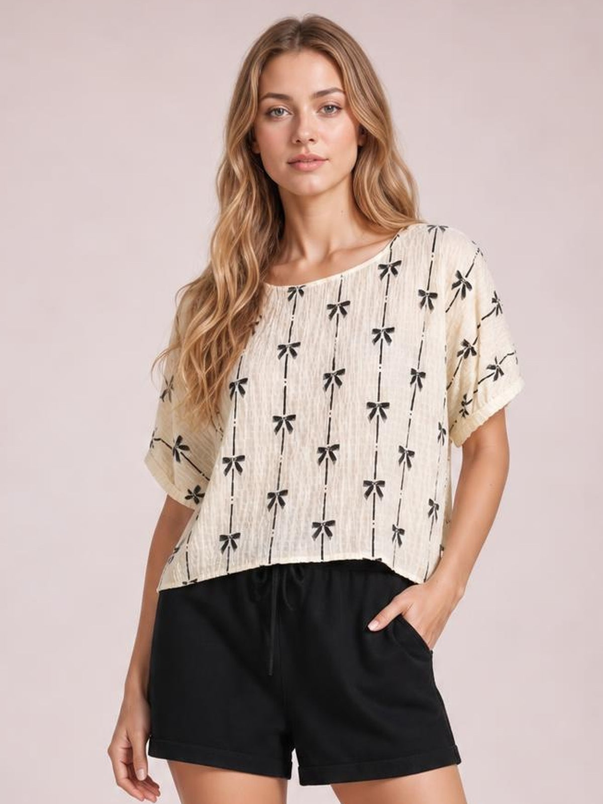 Model poses with one hand in her pocket wearing an ivory short sleeve top with black bow print, a wide neckline, relaxed cropped silhouette, and rolled sleeve cuffs.