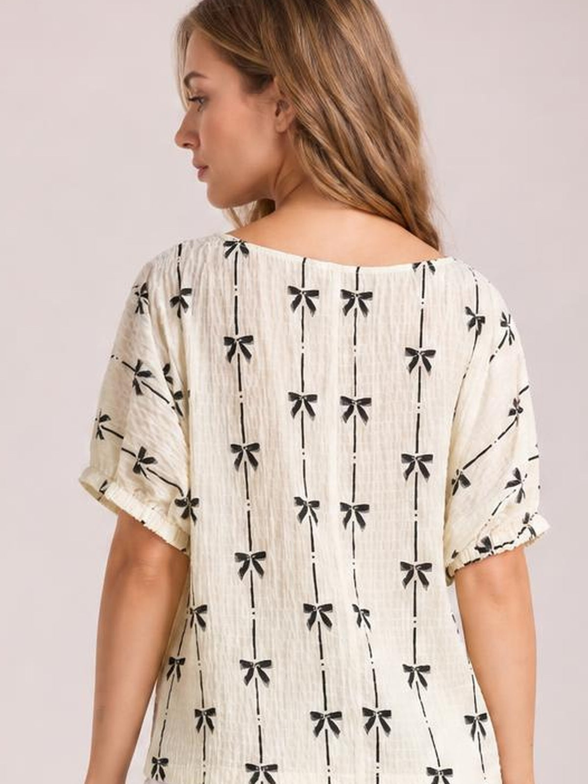 Model shown from the back in a cream Bow Print Short Sleeve Top with black bow-and-line pattern, round neckline, cuffed sleeves, and a relaxed silhouette.