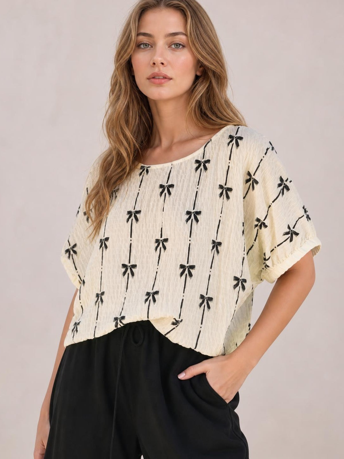 Model wearing an ivory short sleeve top with a black bow print, rounded neckline, loose silhouette, and softly textured fabric, posing with one hand in her pocket.