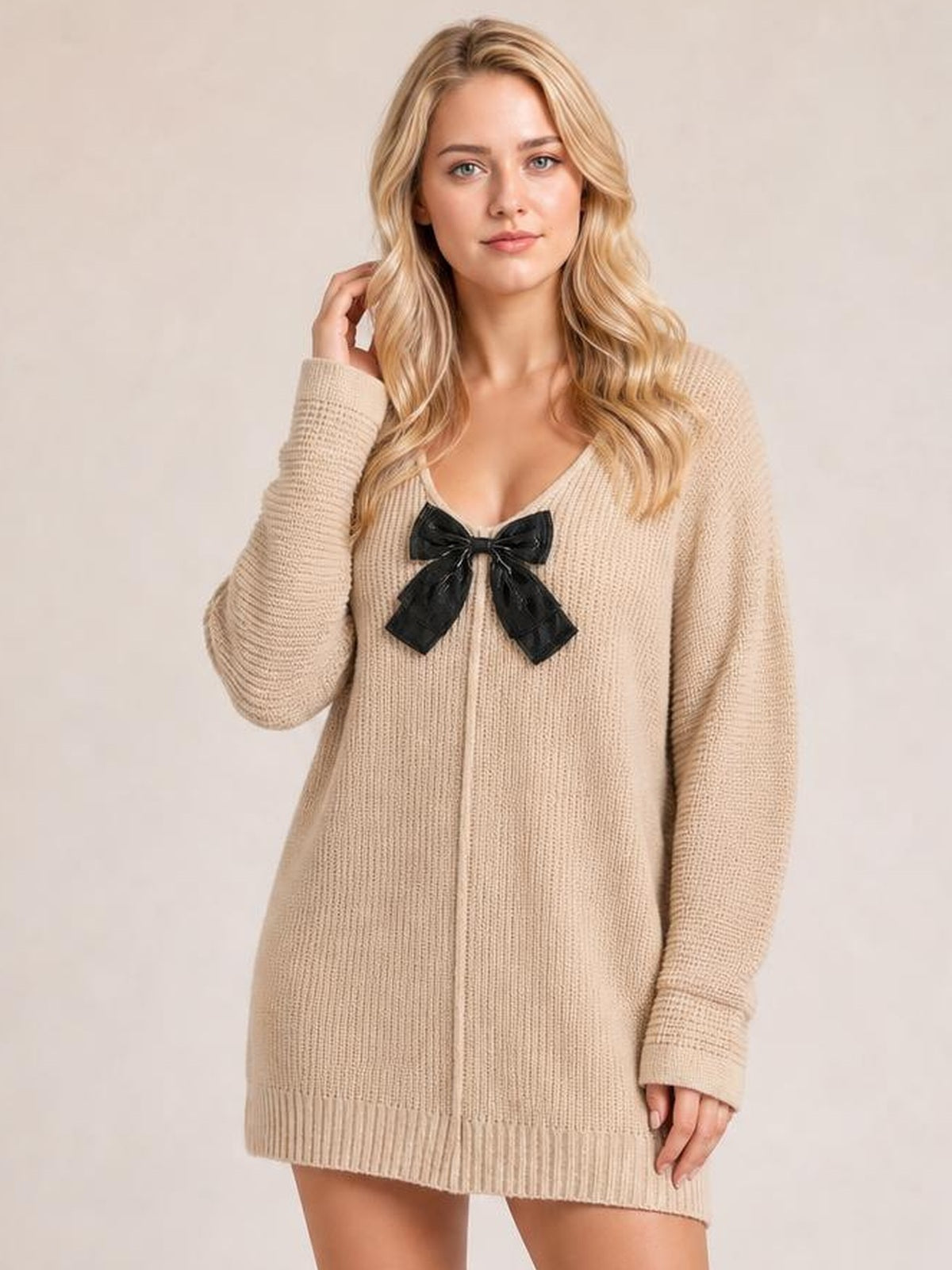 Model wearing a beige ribbed knit sweater dress with long sleeves, a relaxed silhouette, V-neckline, seam detail, and black bow, touching her hair.