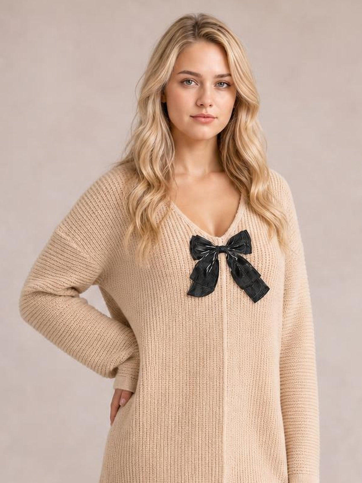 Model poses in a beige ribbed knit sweater dress with long sleeves, a relaxed silhouette, V-neckline, and contrasting black bow detail at the chest.