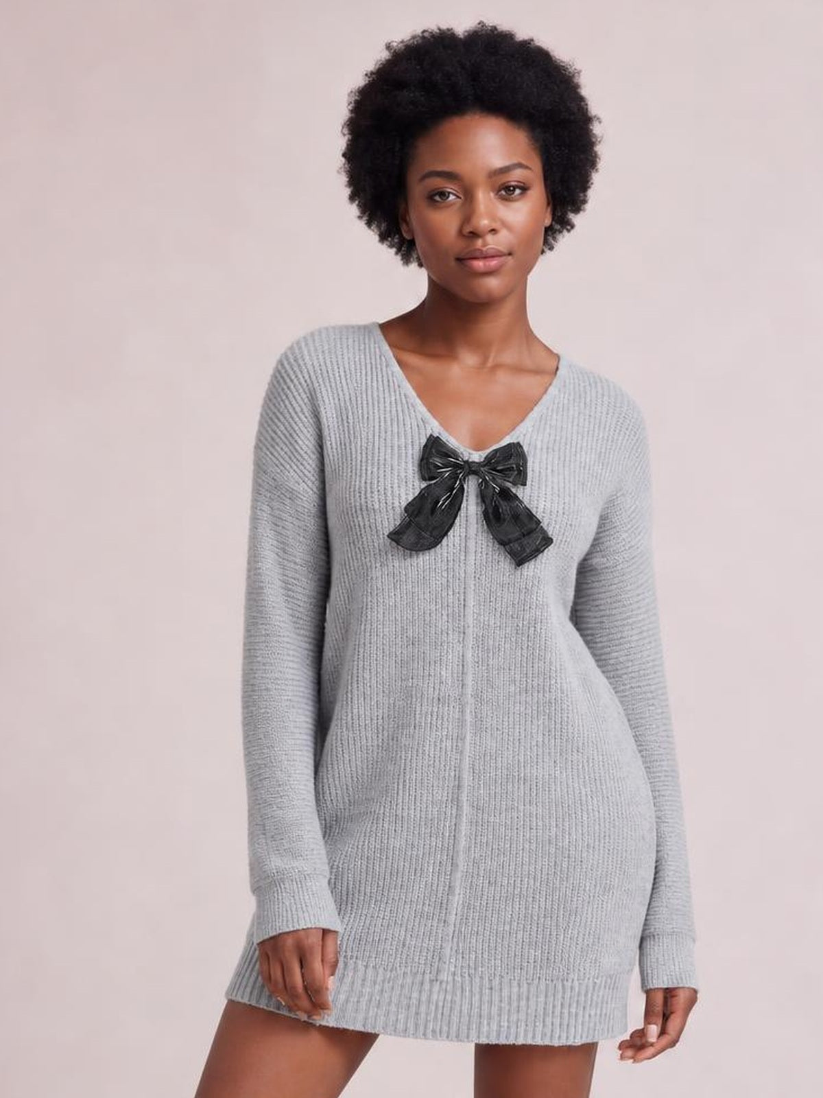Model stands facing forward in a light gray ribbed knit sweater dress with long sleeves, a V-neck, relaxed silhouette, and black bow detail at the chest.