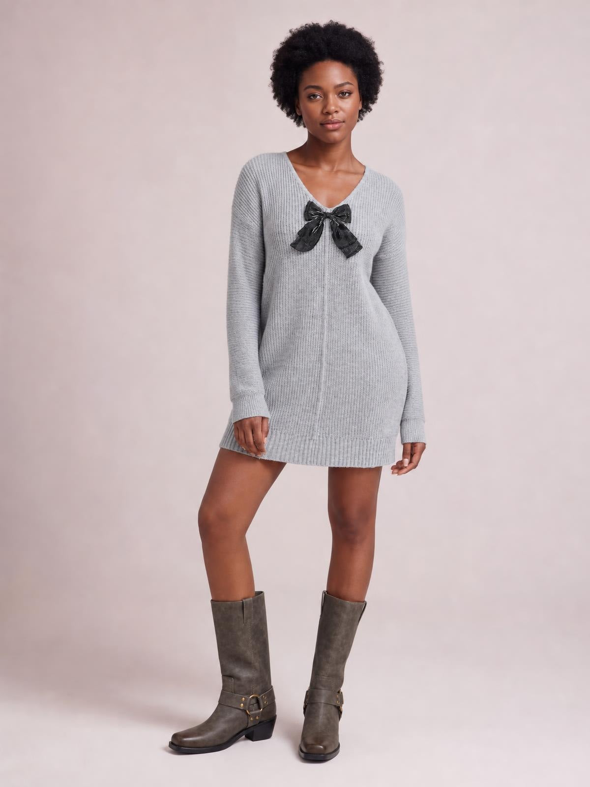 Model poses in a light gray ribbed knit sweater dress with long sleeves, a V-neckline, center seam detail, black bow accent, and relaxed mini silhouette.