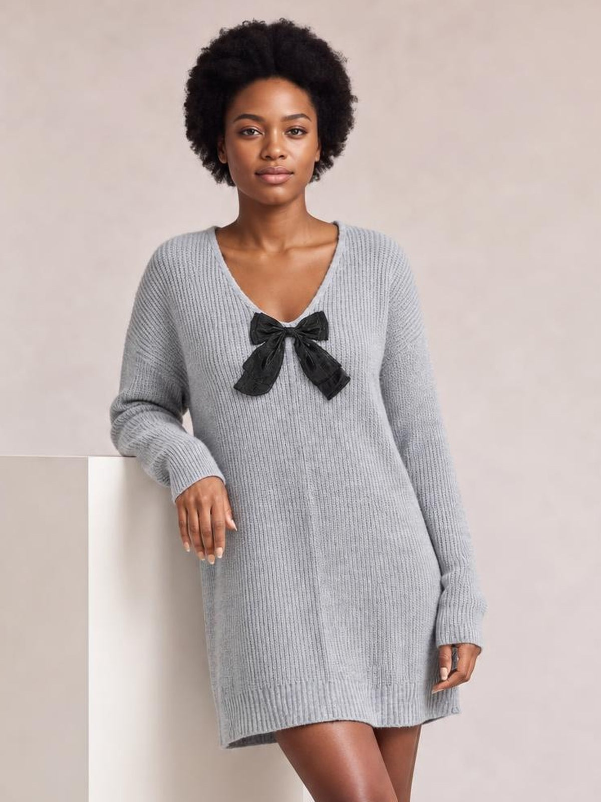 Model poses in a light gray ribbed knit sweater dress with long sleeves, a V-neck, black bow detail, relaxed straight silhouette, and ribbed hem.
