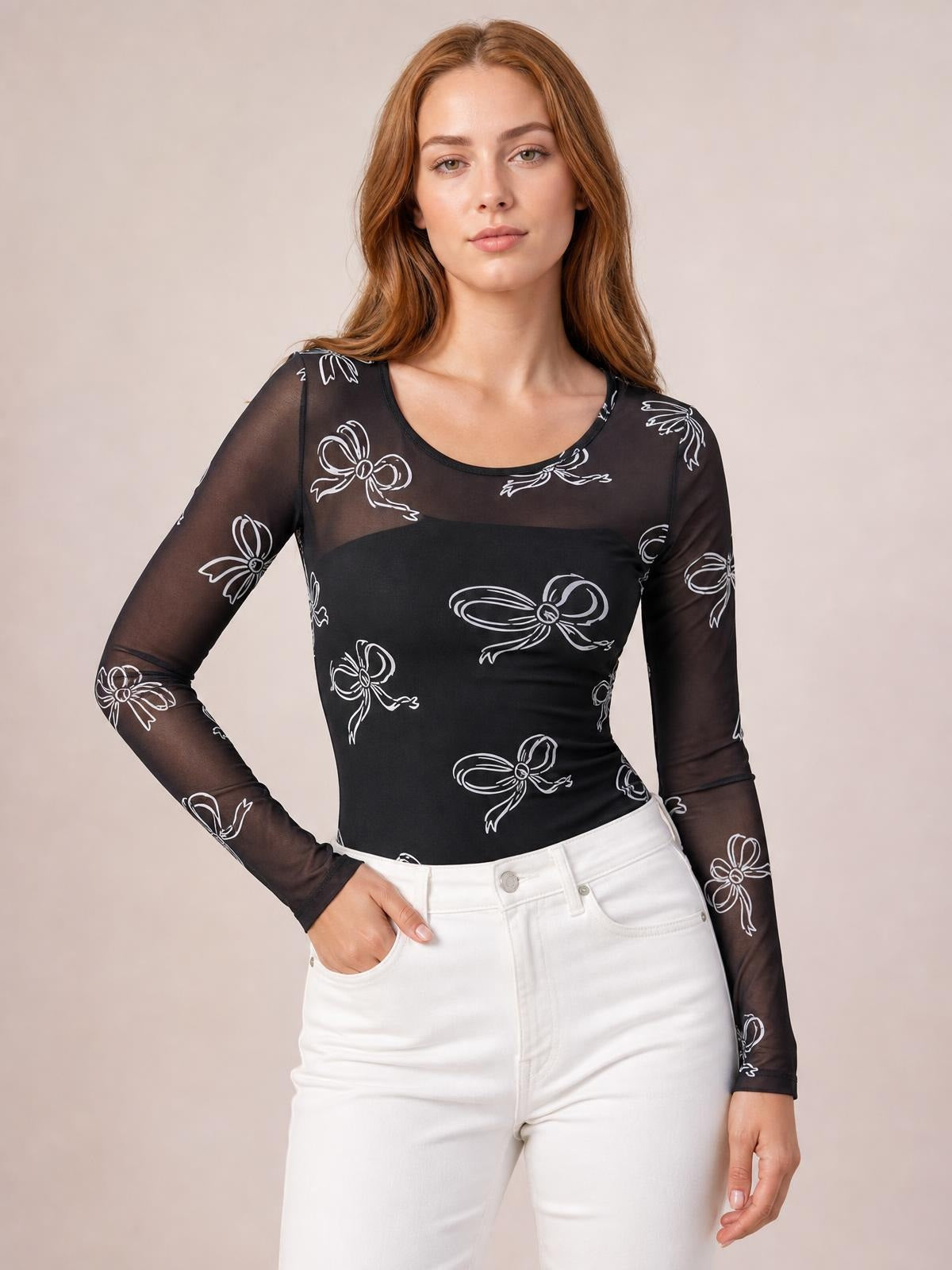 Model poses with one hand in her pocket wearing a black sheer bow-print round-neck top with fitted long sleeves and a slim silhouette.